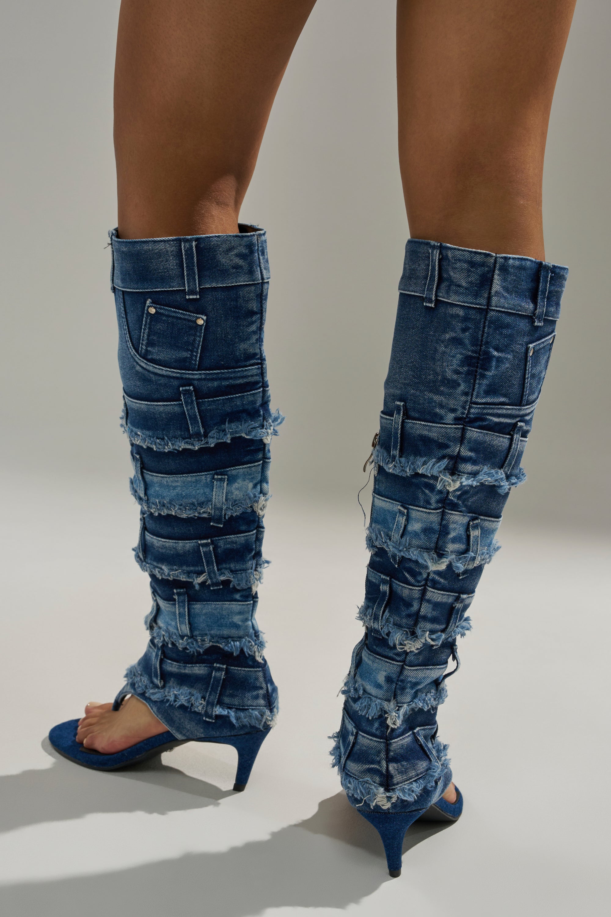 A person wears the AKIRA GOLD MAHONEY DENIM PANT DETAIL GLADIATOR STILETTO—knee-high, open-toe denim boots featuring frayed edges and multiple pockets. The rear shot highlights their unique, distressed style from knee to heel. 3