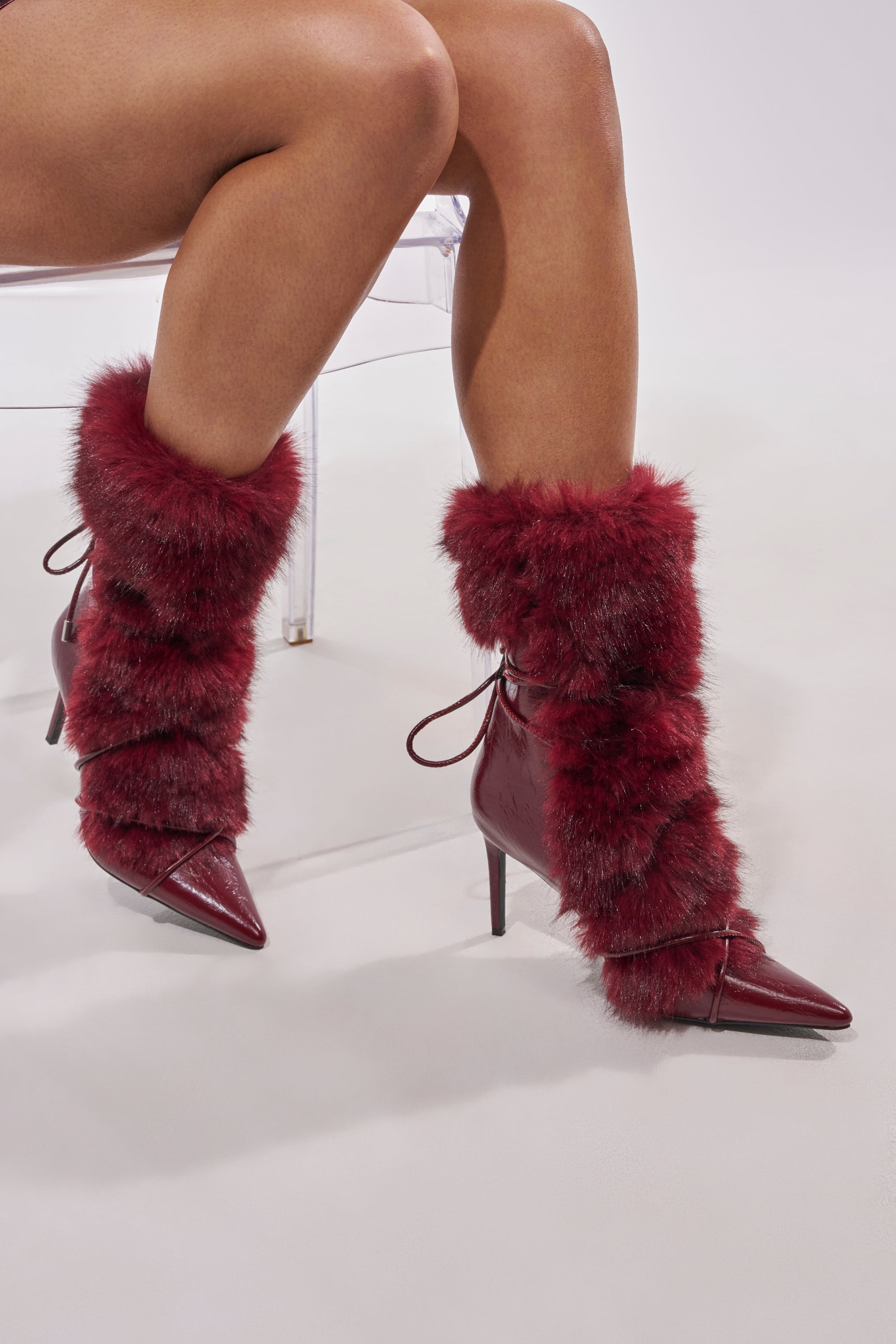A person sits against a plain light background wearing the AZALEA WANG MAHTAMA Burgundy Fur Stiletto Booties, featuring pointed toes and thick, fluffy faux fur trim around the ankles and shins.