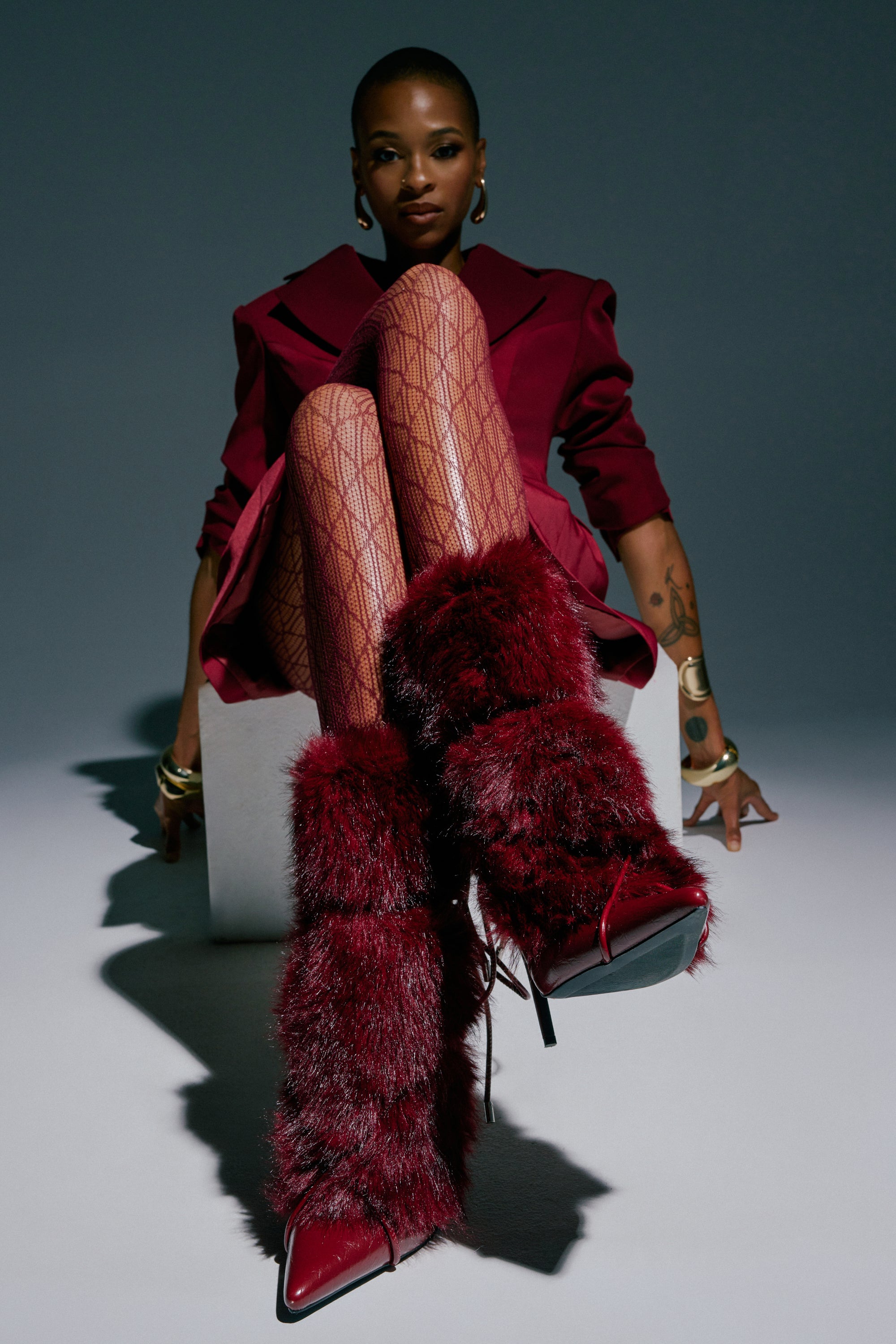 Wearing the AZALEA WANG MAHTAMA BURGUNDY FUR STILETTO BOOTIE, a person in a maroon outfit and patterned tights sits confidently with crossed legs—a bold choice for cold weather style. The gray background and strong shadows accentuate their fashionable pose.