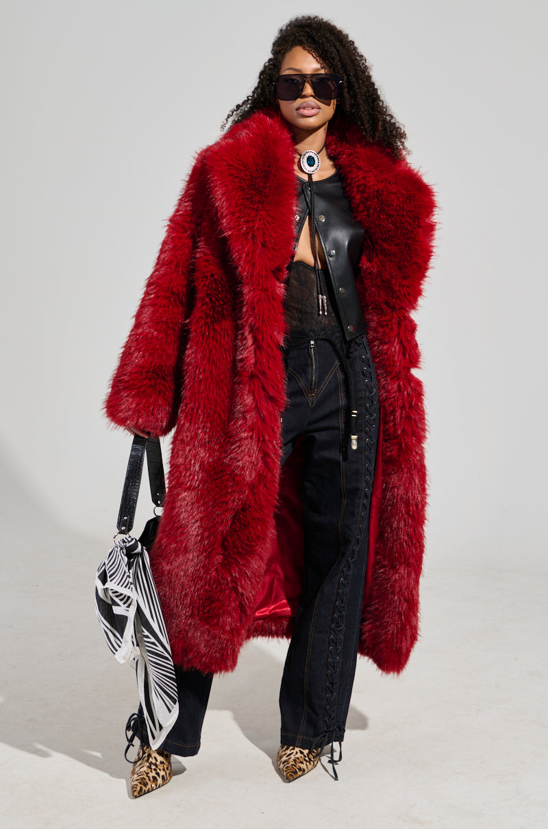 Draped in the luxurious MAJOUR FUR COAT over a sleek black top and pants ensemble, a person radiates confidence. Sporting long curly hair, sunglasses, and leopard-print shoes, they carry an elegant black bag adorned with patterned cloth. The sophisticated look is beautifully highlighted against the plain white background.