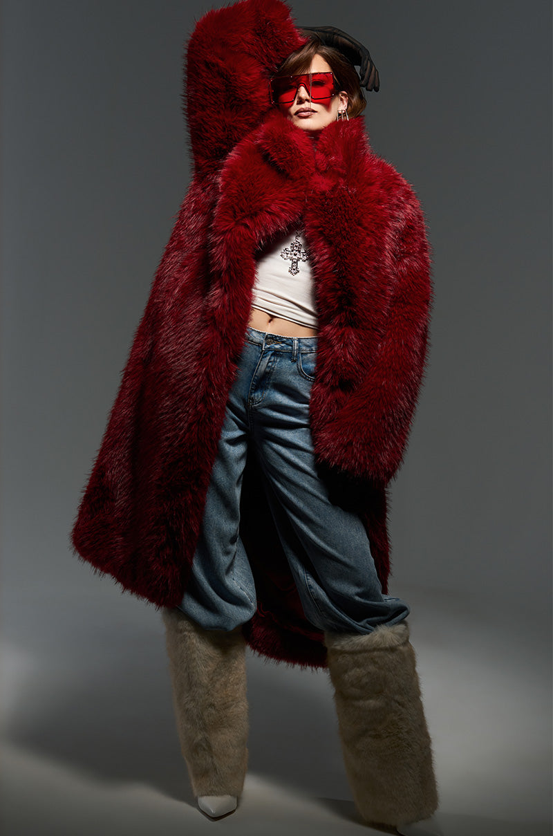 A person wearing the MAJOUR FUR COAT, a white top, and blue jeans poses with one arm raised. They have oversized red sunglasses and are wearing furry boots. The sophisticated finish of their outfit contrasts elegantly against the plain gray background.