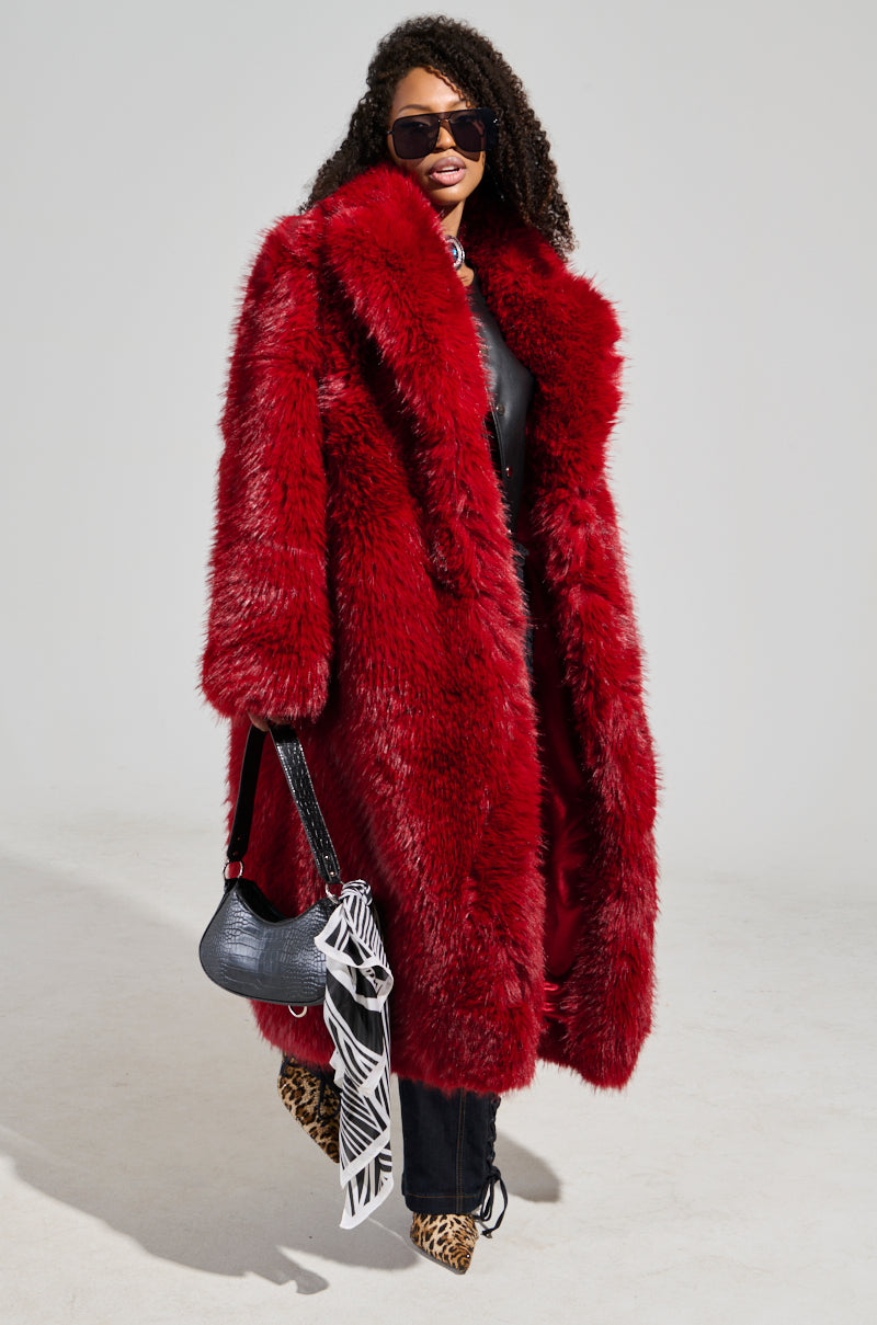 A person with long curly hair wears luxurious outerwear—the MAJOUR FUR COAT in a bold red faux fur—and sunglasses. They hold a black handbag with white details, paired with black pants and leopard print shoes, achieving a sophisticated finish against a plain light background.
