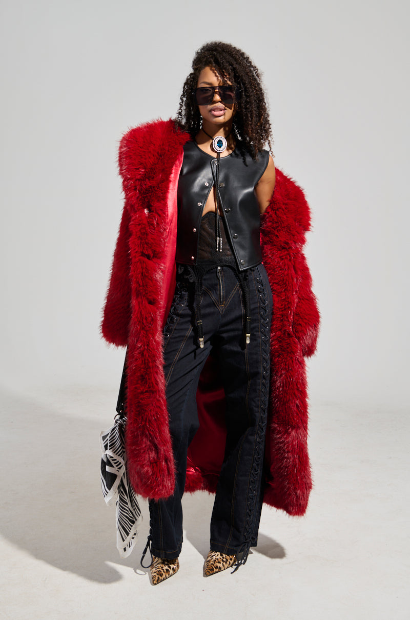 Dressed in the eye-catching MAJOUR FUR COAT in vibrant red, a person wears a black leather vest and lace top beneath it, standing against a plain background. Their ensemble is completed with curly hair, sunglasses, black pants with lace-up details, chic leopard print shoes, and they are holding an elegant black and white striped bag.