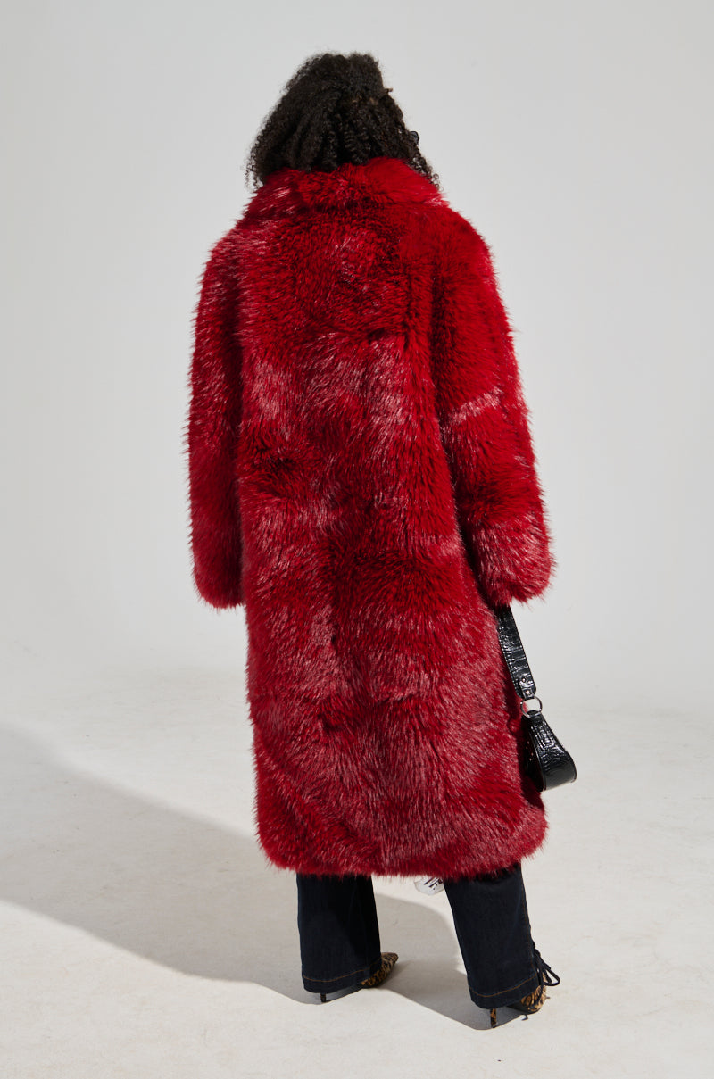 A person with curly hair is donning the MAJOUR FUR COAT in red, paired with dark pants and carrying a black purse. The sophisticated finish of the ensemble stands out against the plain, light-colored background as the individual faces away from the camera.