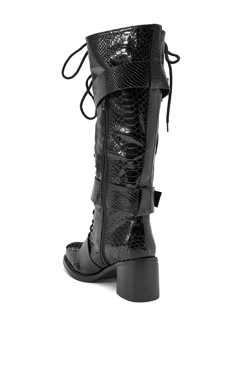 The AZALEA WANG MAKAR BLACK CROC LACE UP DETAIL BOOT is a knee-high black croc boot with a chunky heel, shiny faux snakeskin texture, lace-up detail, and decorative side flaps.