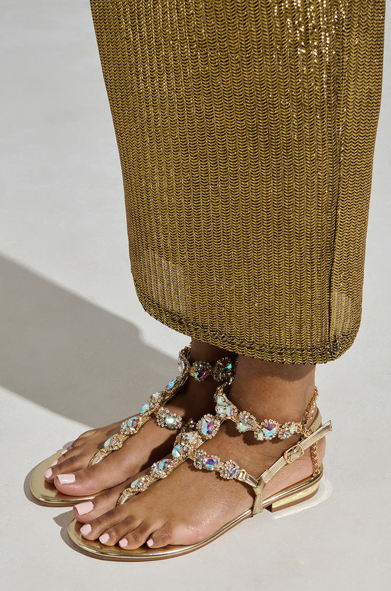 A person wearing AZALEA WANG MANAIA GOLD EMBELLISHED SANDALS stands on a beige surface. The metallic gold base perfectly matches their shimmering, ankle-length skirt or dress. Their pale pink toenails add an elegant touch to the stylish look.
