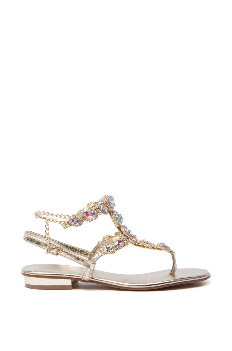 The AZALEA WANG MANAIA GOLD EMBELLISHED SANDAL features a side view of a metallic gold PU design with intricate beadwork, rhinestones, multi-colored gemstones, thin straps, and a flat sole with a slight heel for an elegant yet comfortable look.