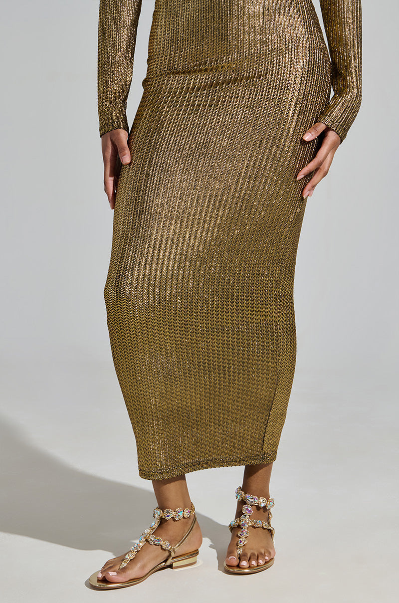 A person stands gracefully in a long, shimmering metallic gold dress with a ribbed texture, their hands gently resting on the fabric. The AZALEA WANG MANAIA GOLD EMBELLISHED SANDAL sparkles beneath, adding elegance against the neutral background.