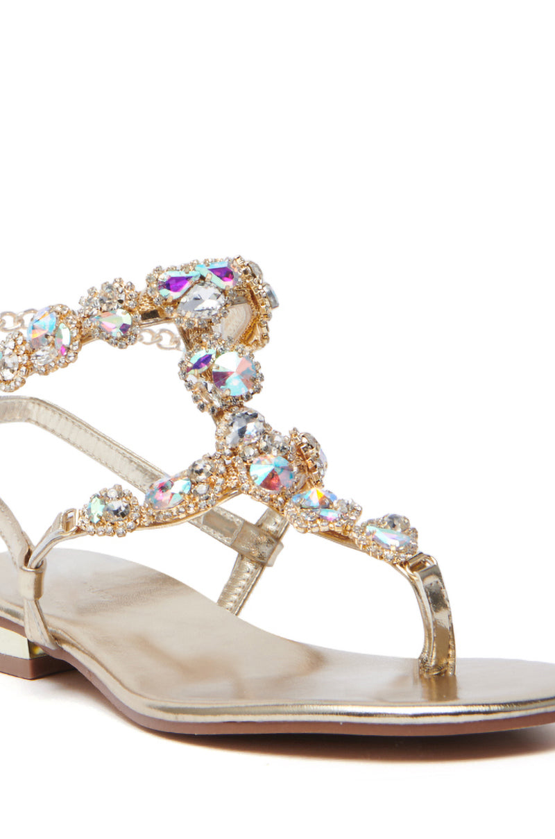 The AZALEA WANG MANAIA GOLD EMBELLISHED SANDAL features a thong design with intricate rhinestone-adorned straps, reflecting multiple colors. Its chain link ankle strap adds elegance to the flat sole, contributing a glamorous flair.
