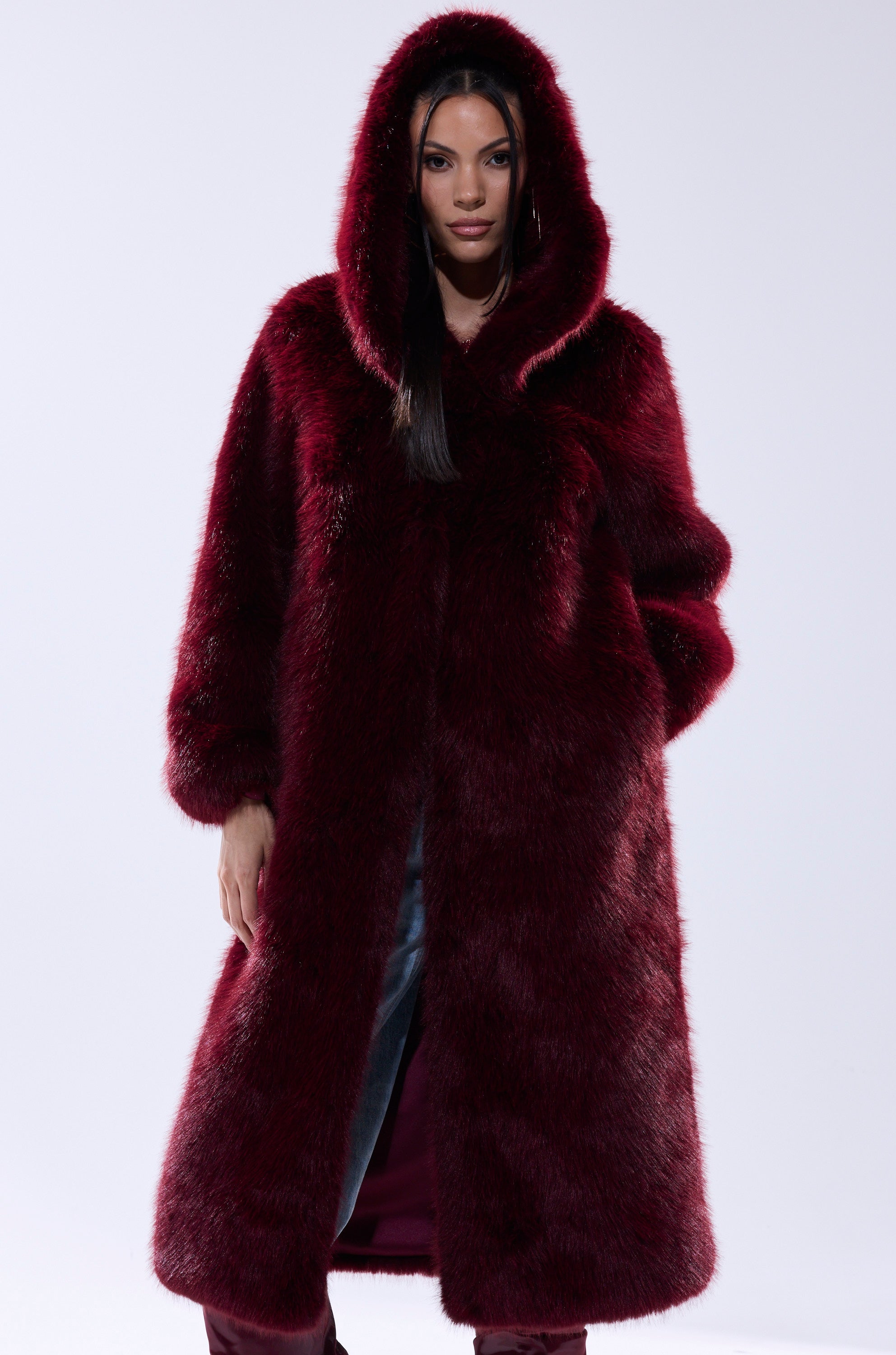 A woman stands facing forward against a white background, wearing the MANCHESTER HOODED FAUX FUR IN BURGUNDY. Her dark hair peeks from beneath the hood and her hands rest by her sides.