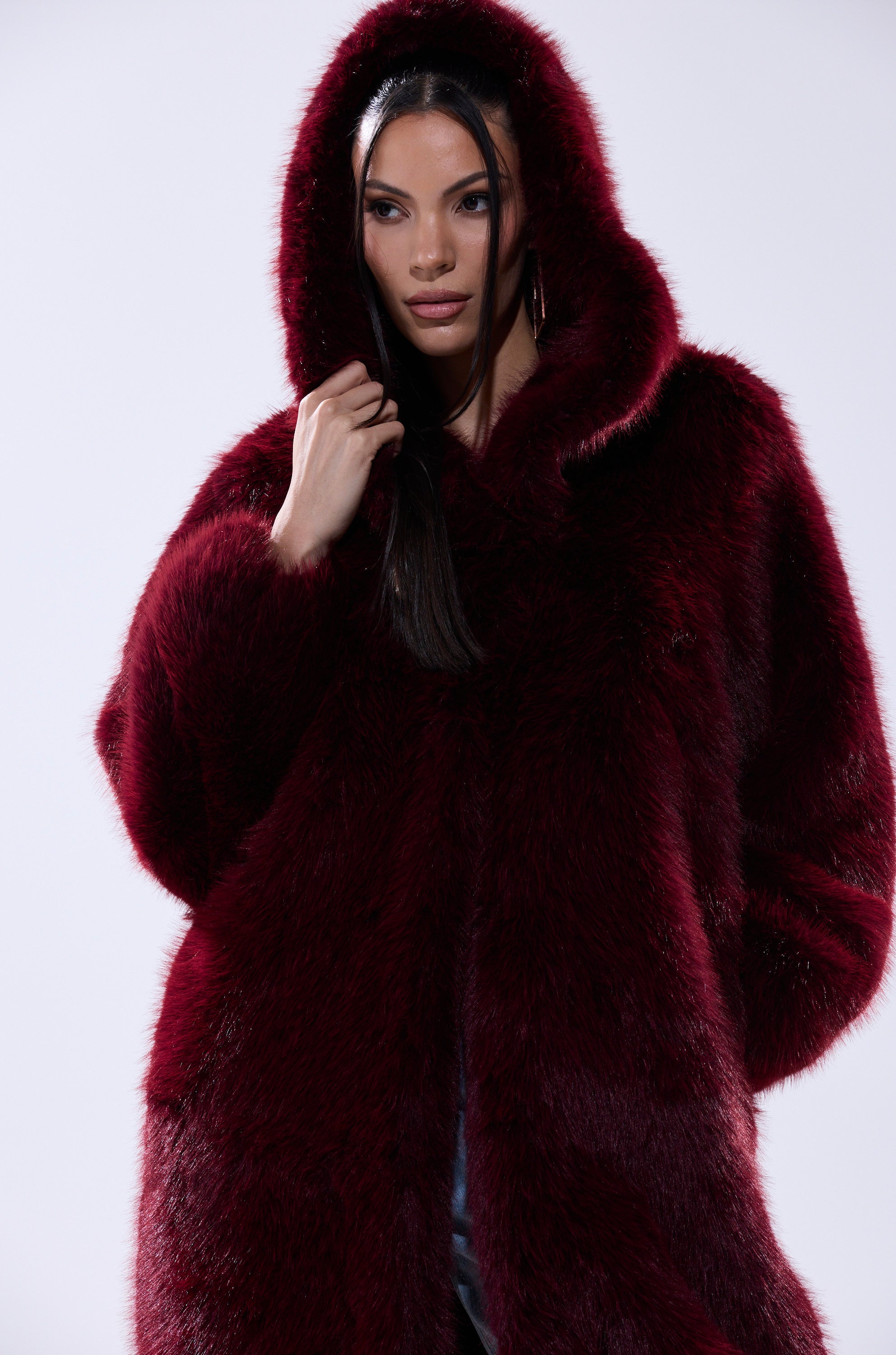 A woman with long dark hair wears the MANCHESTER HOODED FAUX FUR IN BURGUNDY, posing against a plain light background. She gazes slightly to the side with a neutral expression.