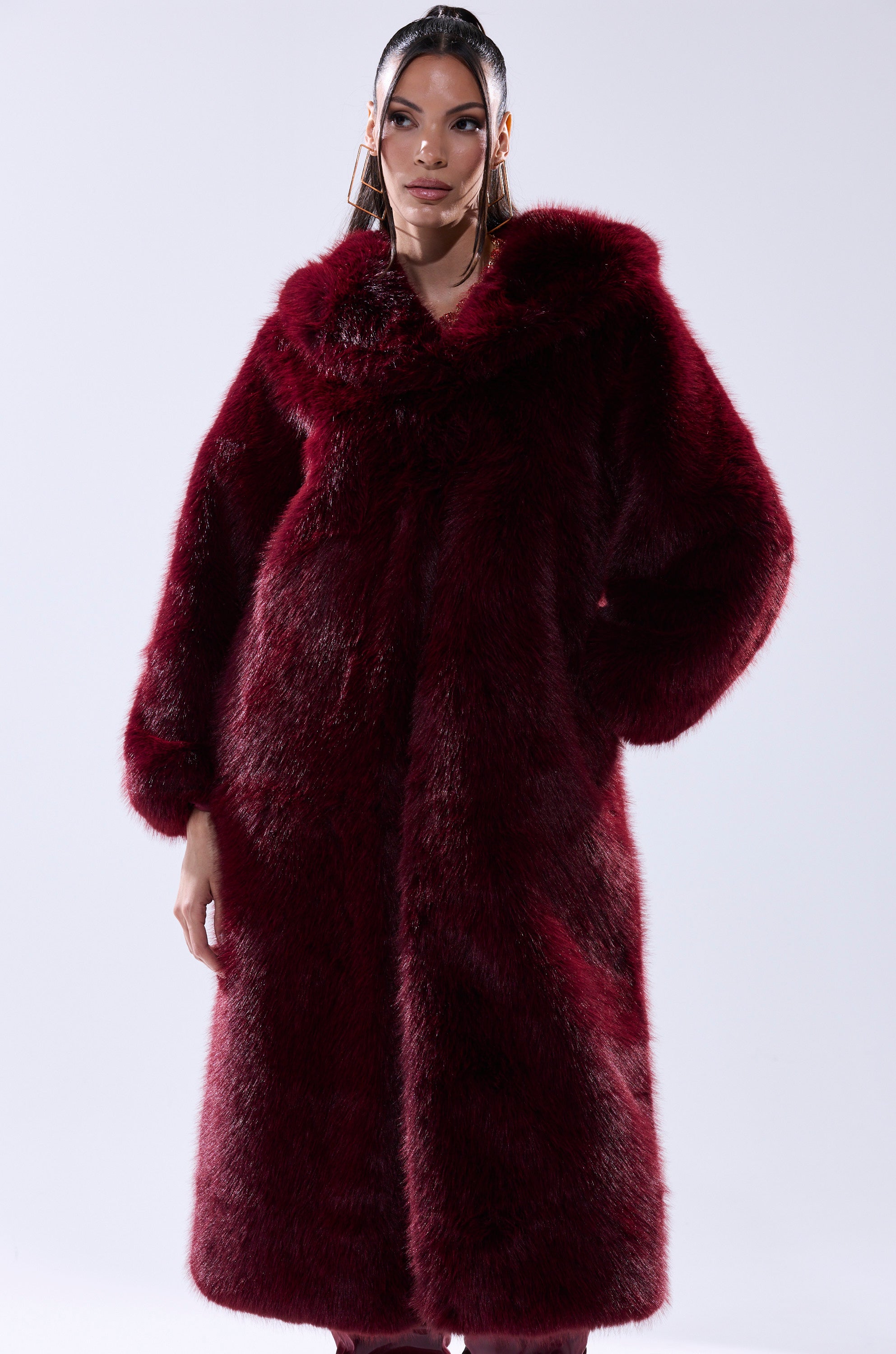 A woman with slicked-back hair stands confidently, hand on hip, wearing the MANCHESTER HOODED FAUX FUR IN BURGUNDY against a plain light background.