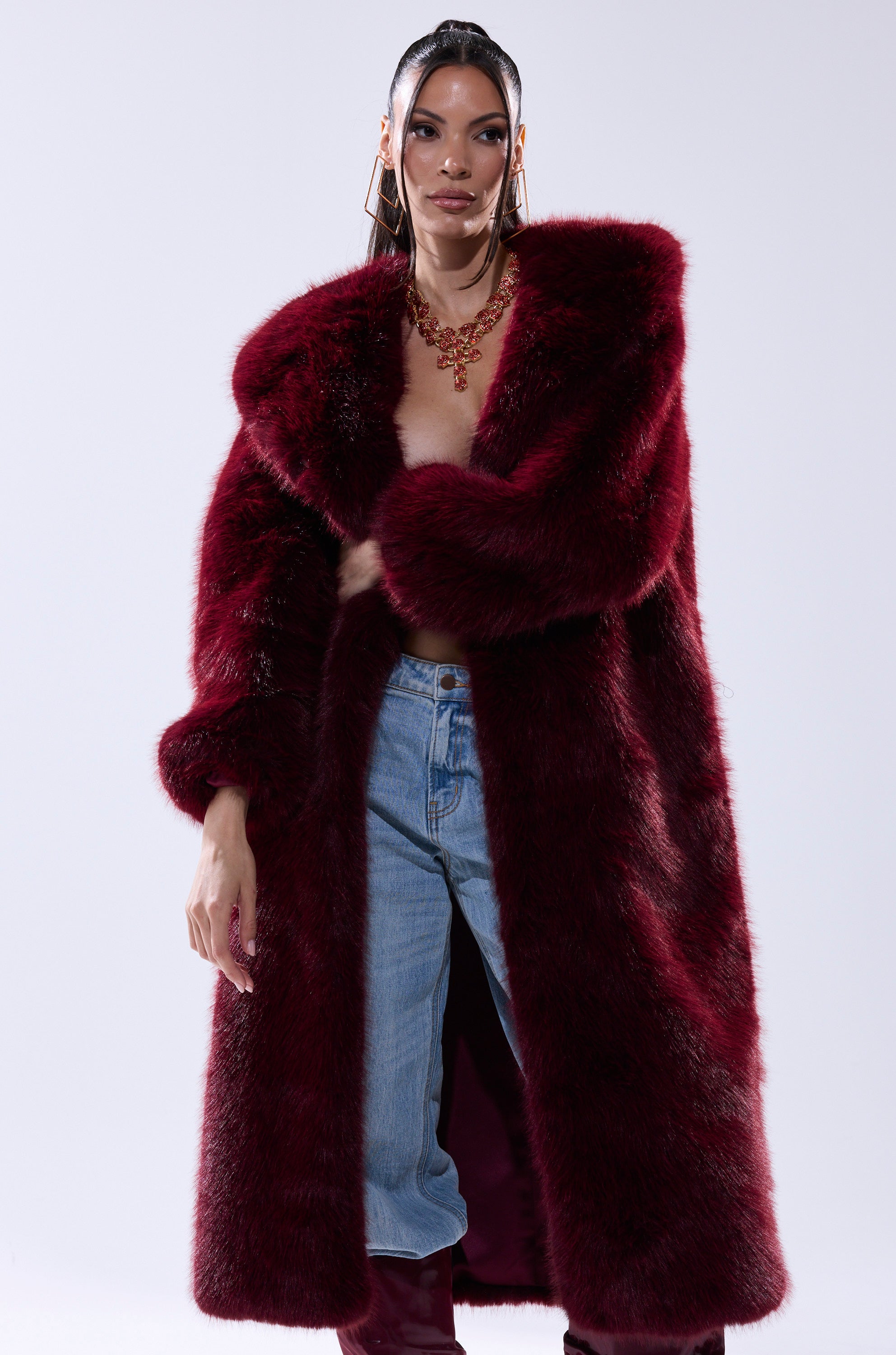 A woman poses in the MANCHESTER HOODED FAUX FUR IN BURGUNDY, styled open over blue jeans and a statement necklace. She sports sleek, wet-styled hair and large hoop earrings against a simple light background.