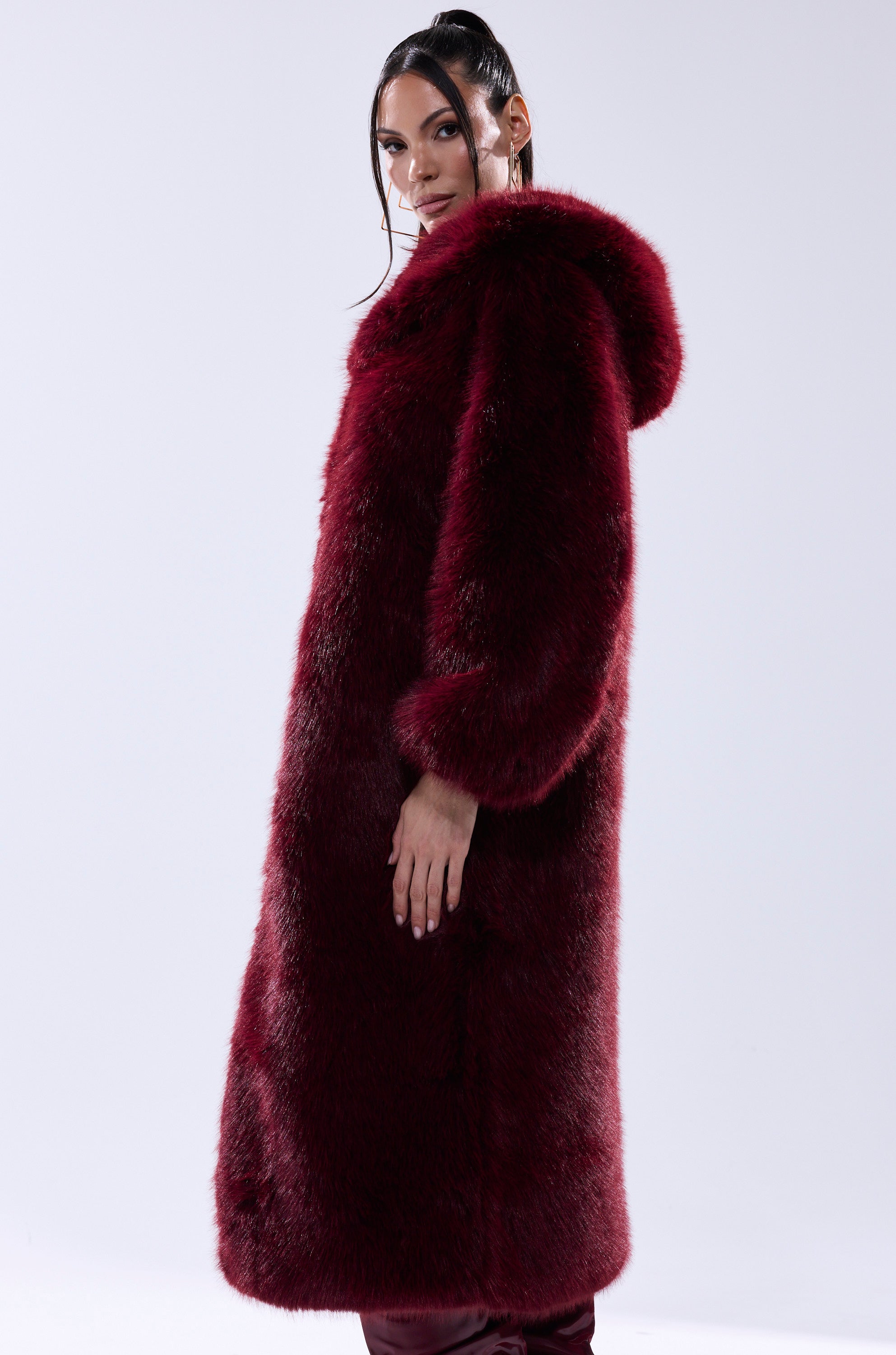 A person with long dark hair stands sideways against a plain light background, wearing the MANCHESTER HOODED FAUX FUR IN BURGUNDY.