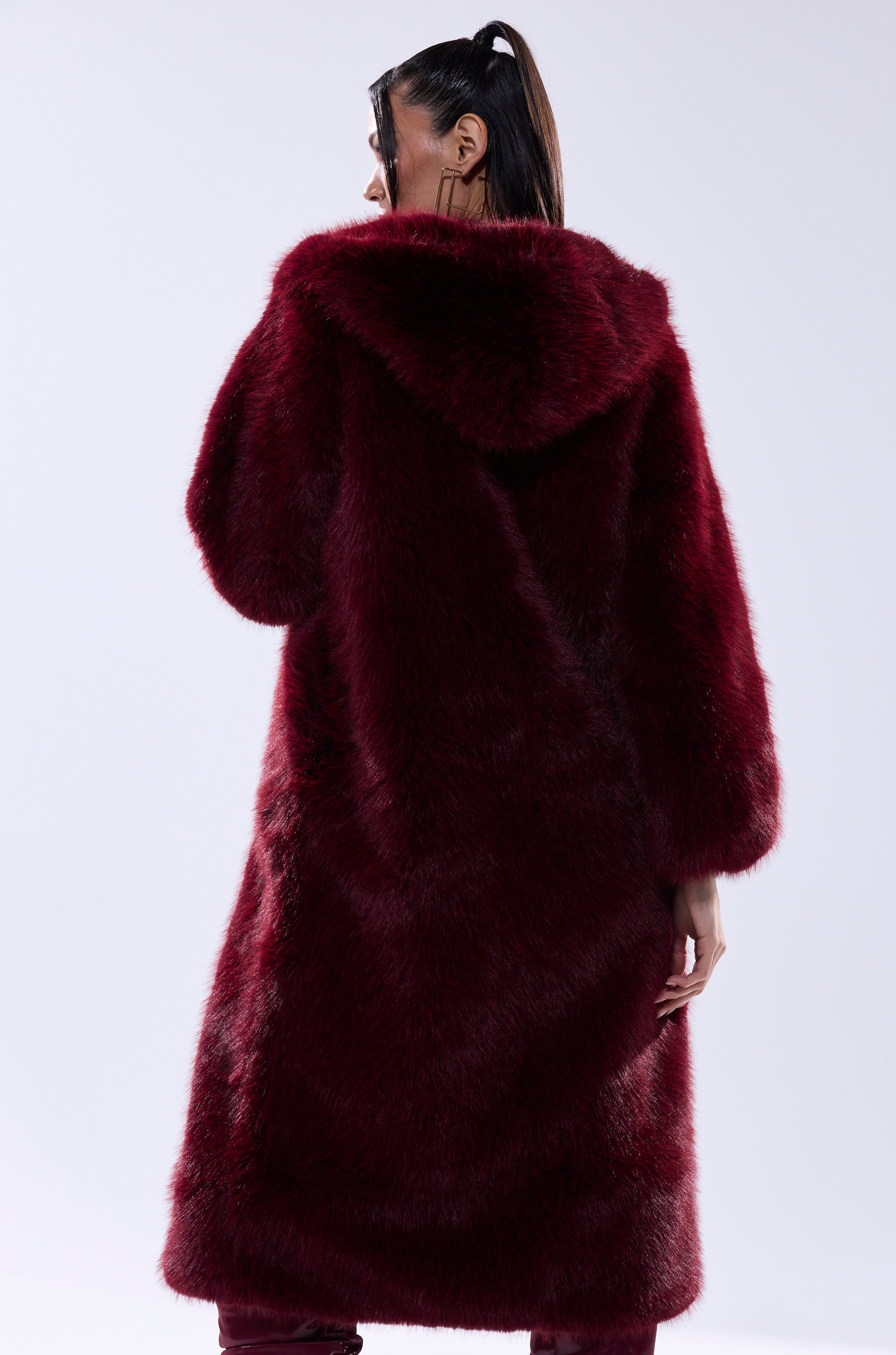 A person with long dark hair in a ponytail is seen from behind, wearing the MANCHESTER HOODED FAUX FUR IN BURGUNDY—a luxurious deep red jacket. The background is plain and light-colored.