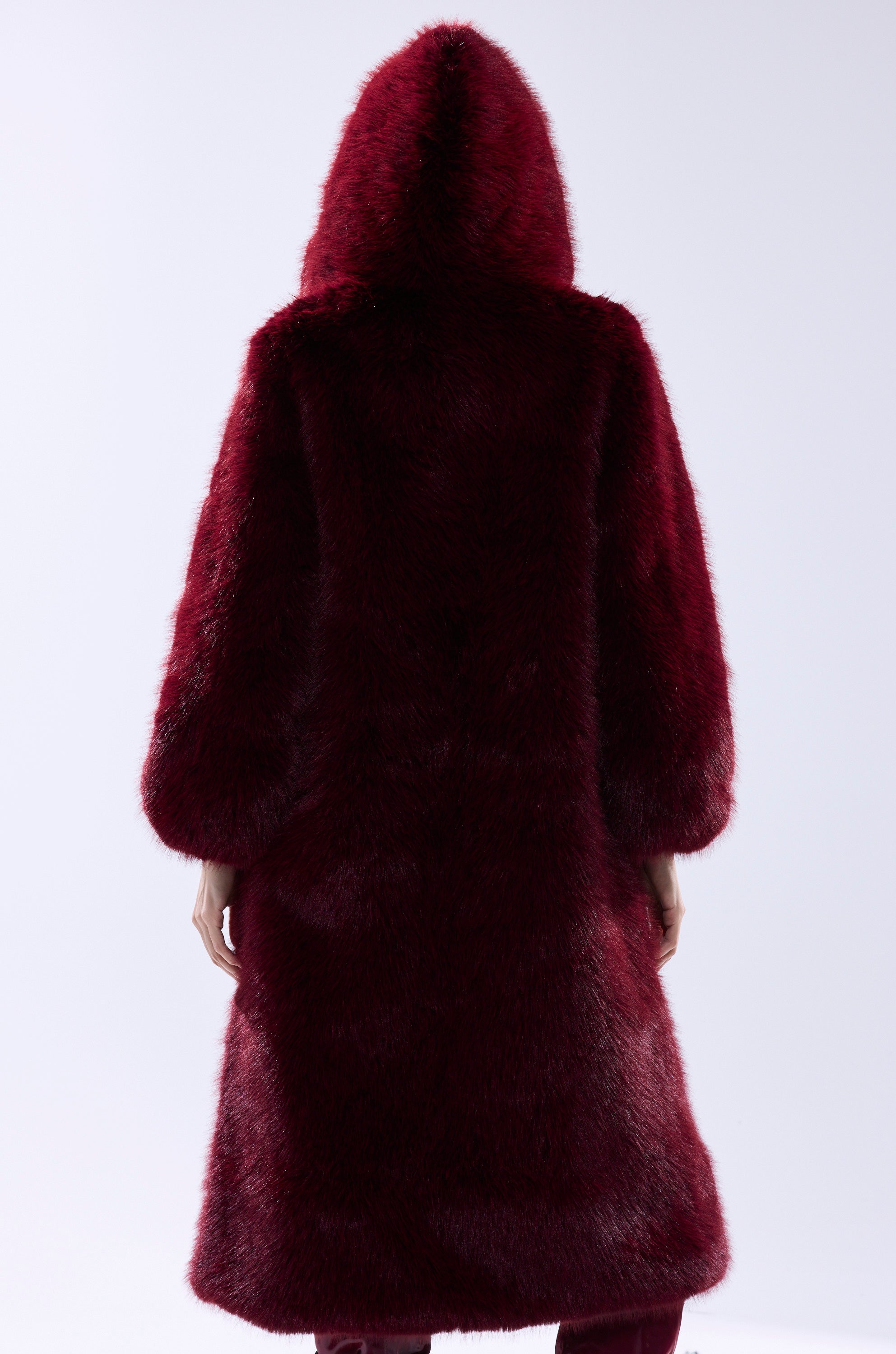 A person is shown from behind wearing the MANCHESTER HOODED FAUX FUR IN BURGUNDY, a long, hooded faux fur coat, set against a plain light background.