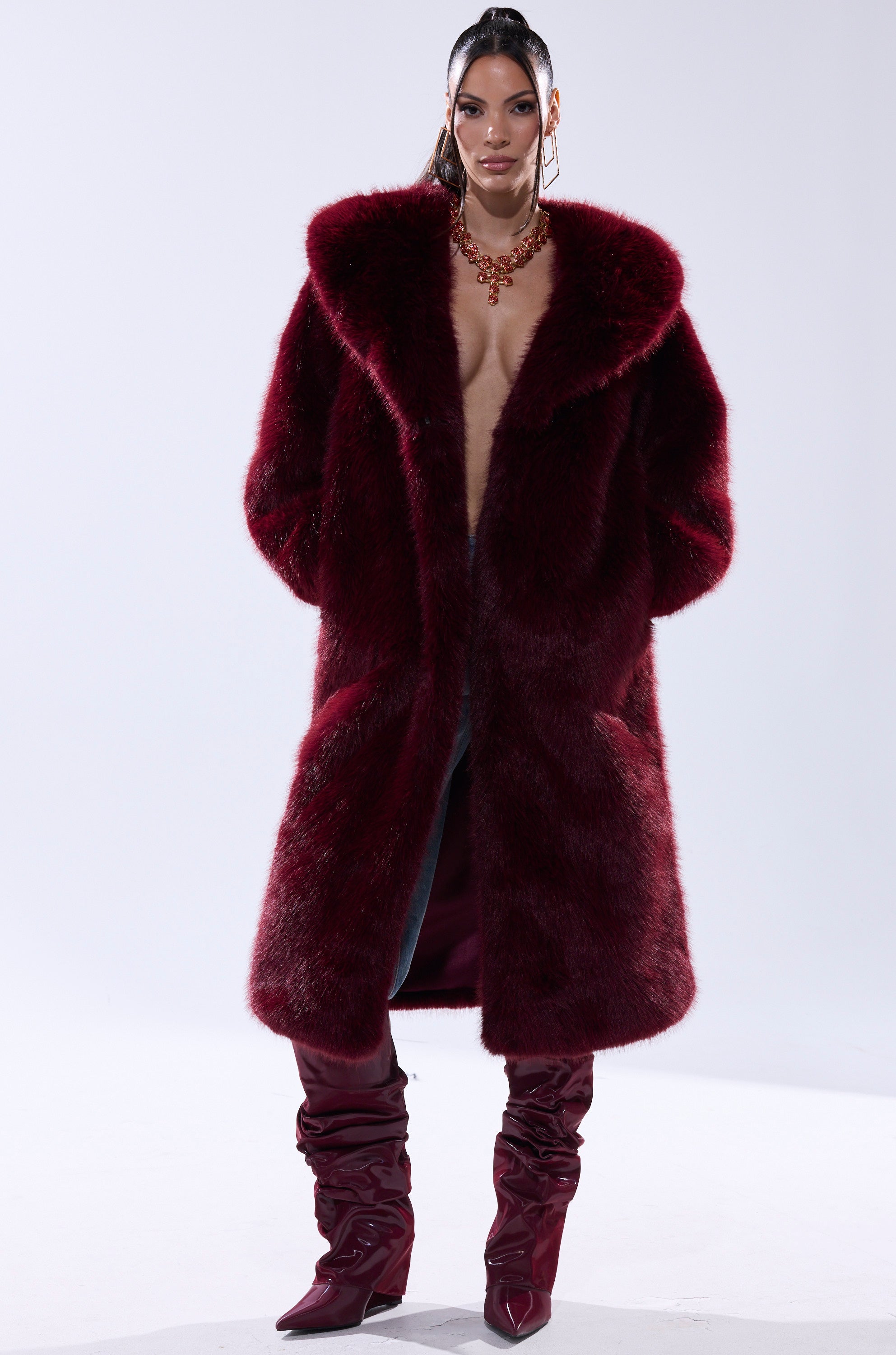 A woman models the MANCHESTER HOODED FAUX FUR IN BURGUNDY—a long, deep red jacket with a wide collar—paired with matching knee-high boots and a statement necklace, her hair slicked back against a plain white background.