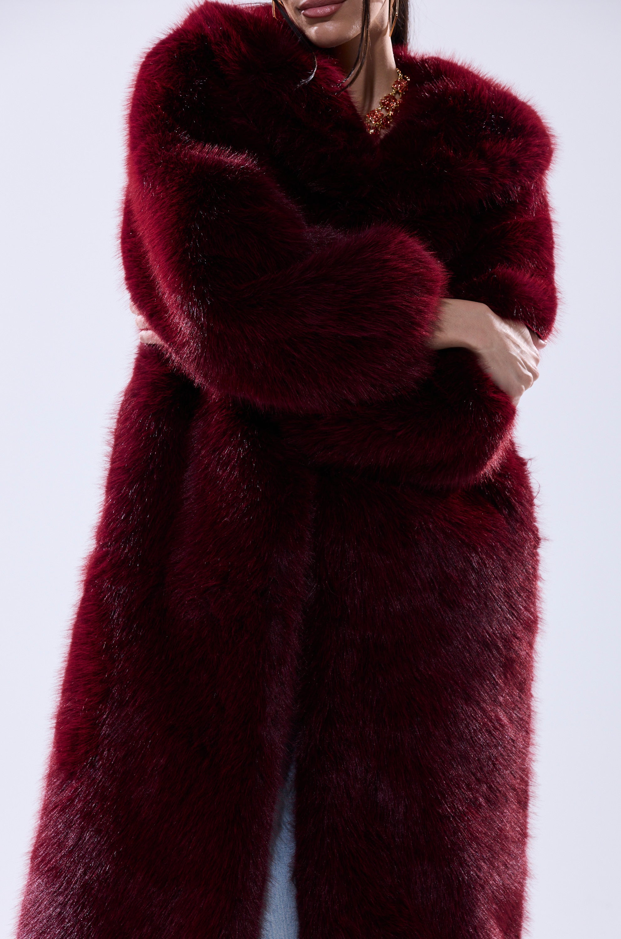 A woman models the MANCHESTER HOODED FAUX FUR IN BURGUNDY, standing against a plain light background with her arms crossed and only her lower face visible.
