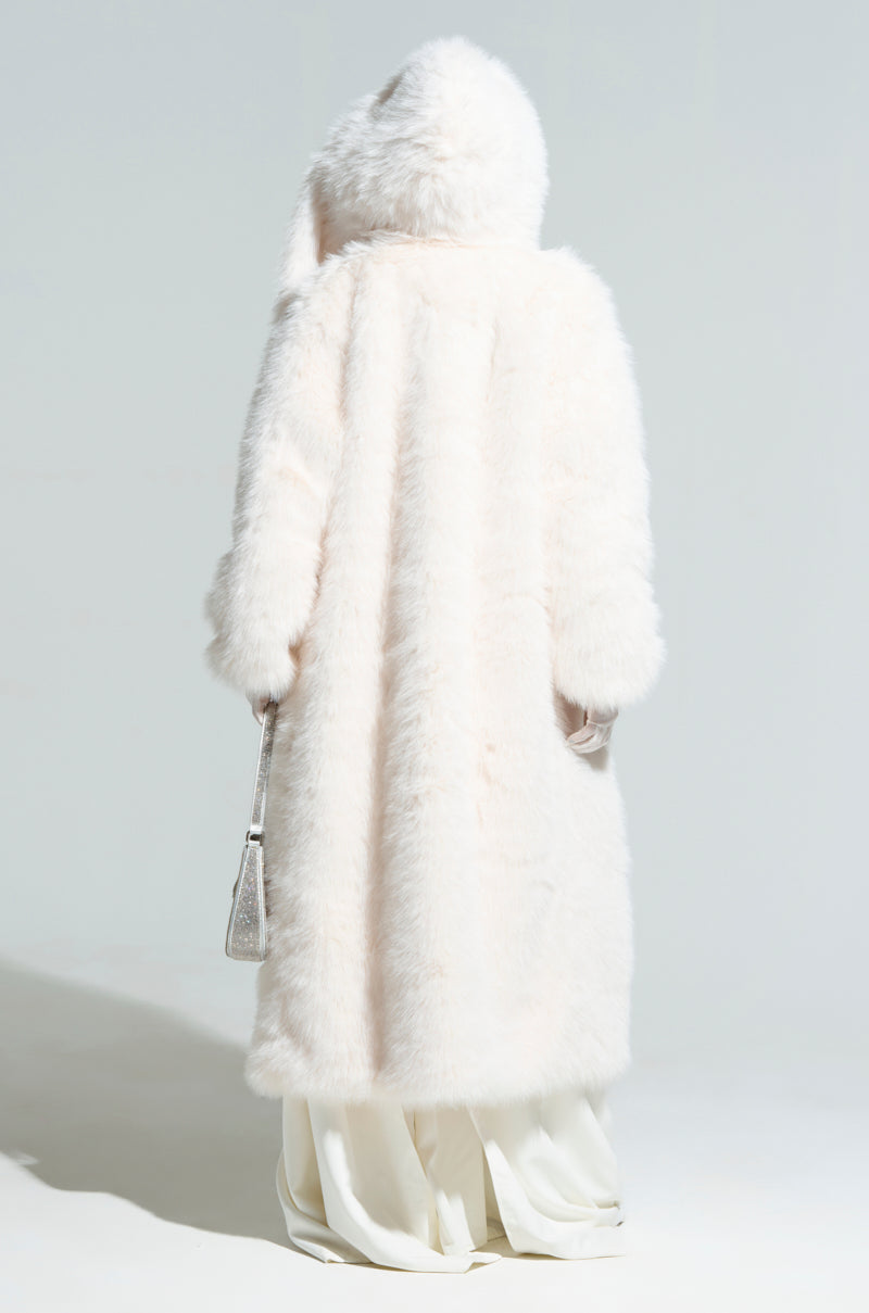 A person in the MANCHESTER HOODED FAUX FUR IN IVORY stands with their back to the camera. They hold a small silver handbag and wear a long white skirt, set against a simple light-colored background.