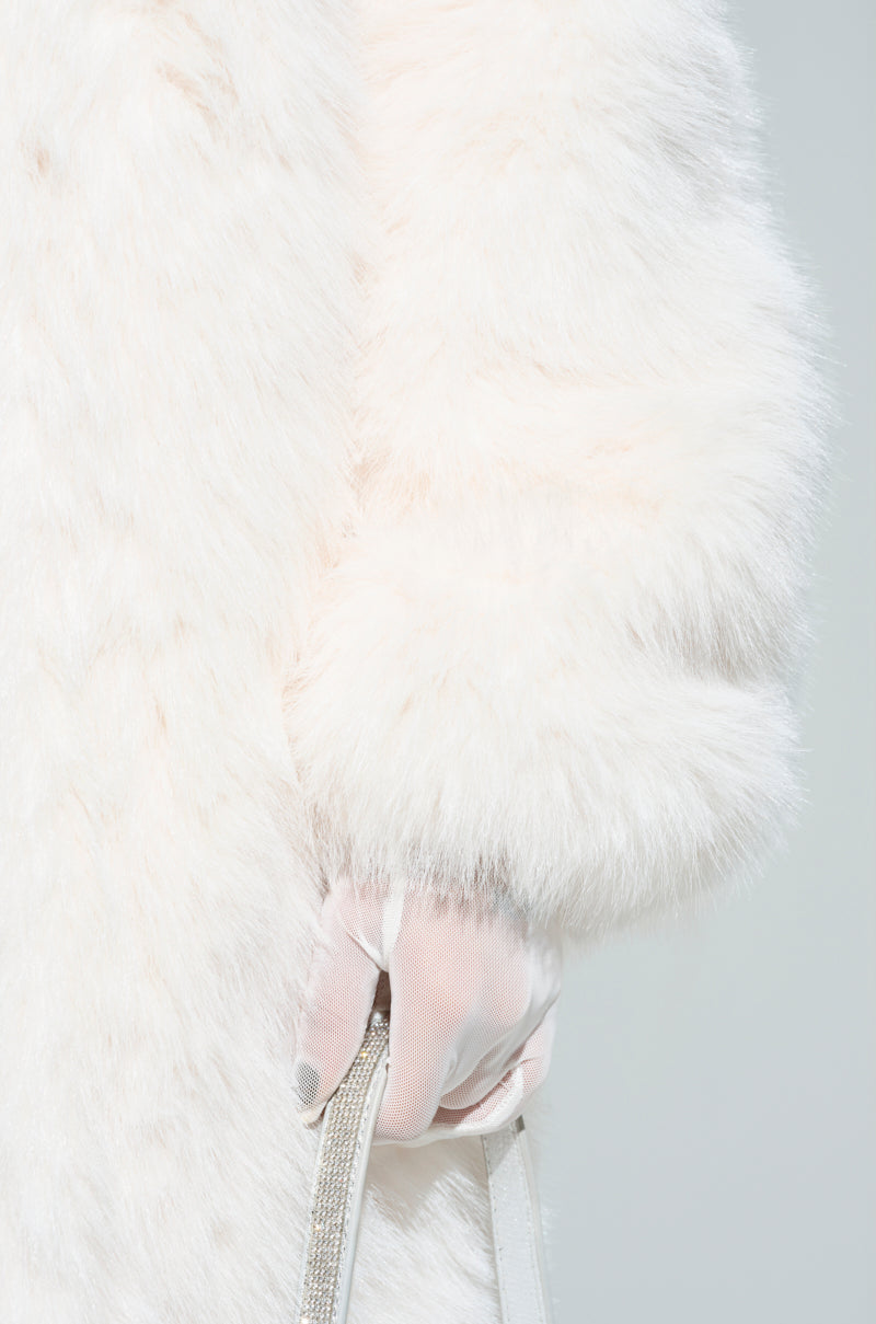 A close-up of someone wearing the MANCHESTER HOODED FAUX FUR IN IVORY, holding a white handbag with a sparkly strap. The light pink fabric glove harmonizes beautifully with the coat's texture against a soft light grey background.