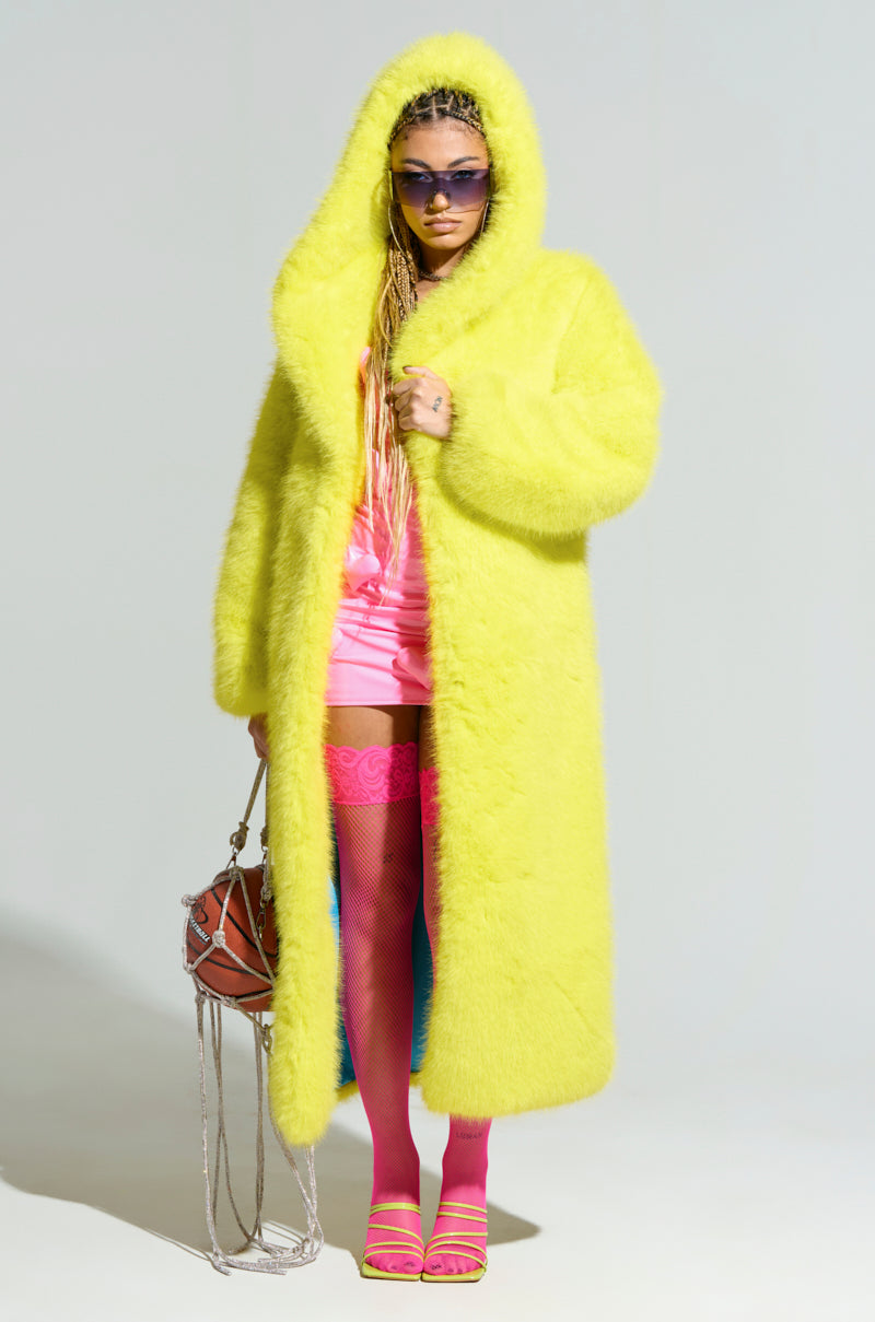 A person wearing the Manchester Hooded Faux Fur in Lime paired with oversized purple sunglasses, pink shorts, and pink thigh-high stockings holds a brown handbag with chain straps and sports yellow sandals. The plain background complements their bold outfit.