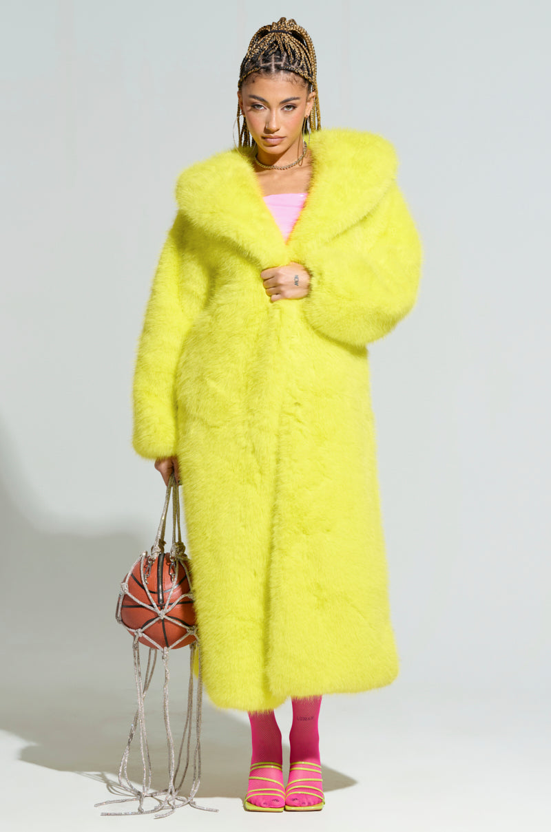 A person stands confidently wearing the Manchester Hooded Faux Fur in Lime, paired with a pink top and pink shoes, their hair styled in colorful braids. They hold a spherical bag with tassels against a plain light gray background.