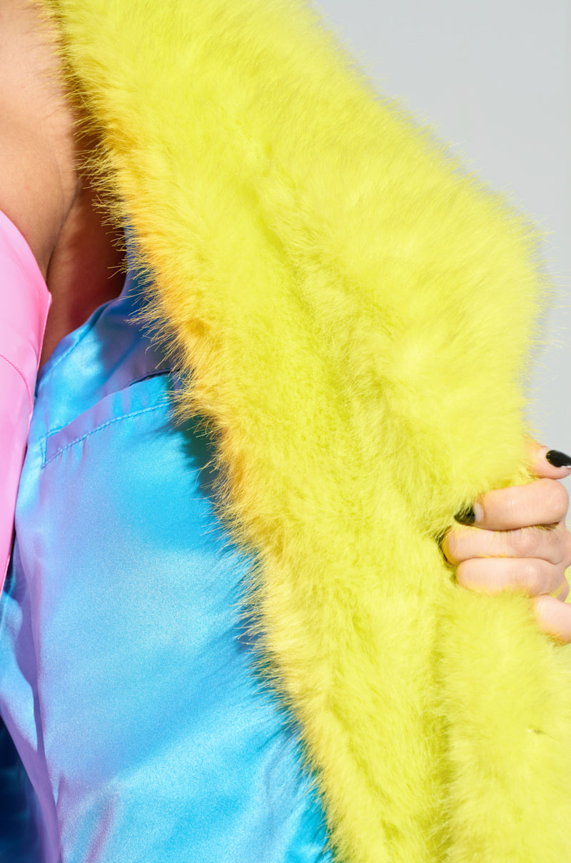 A person holds the Manchester Hooded Faux Fur in Lime against a colorful outfit with glossy blue and pink fabric, highlighting the texture and vibrancy of the materials and showcasing a bold and fashionable style.