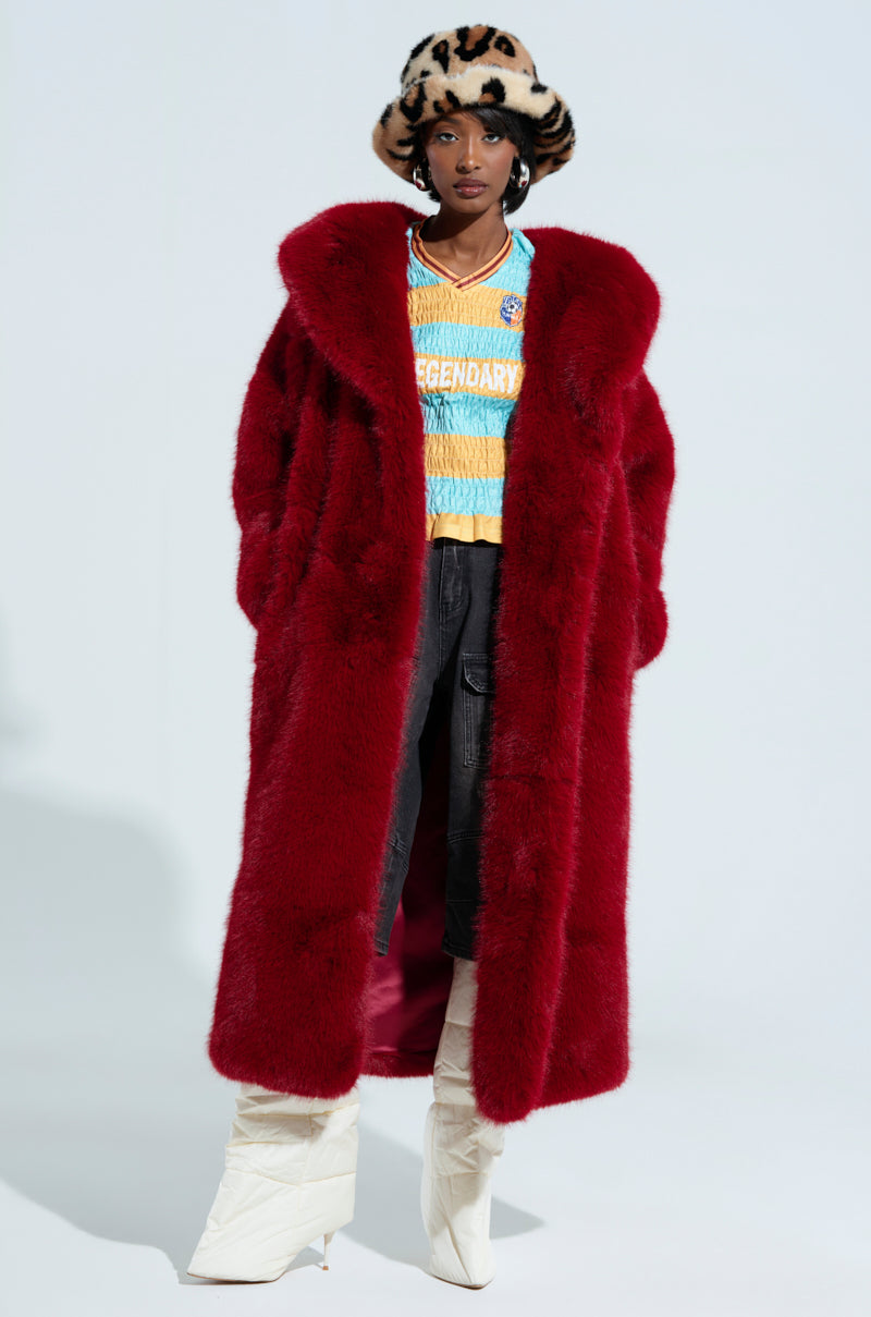 A person wearing the opulent Manchester Hooded Faux Fur in Wine, a vibrant "Legendary" sweater, dark pants, white quilted boots, and a leopard-print hat poses confidently against a plain background.