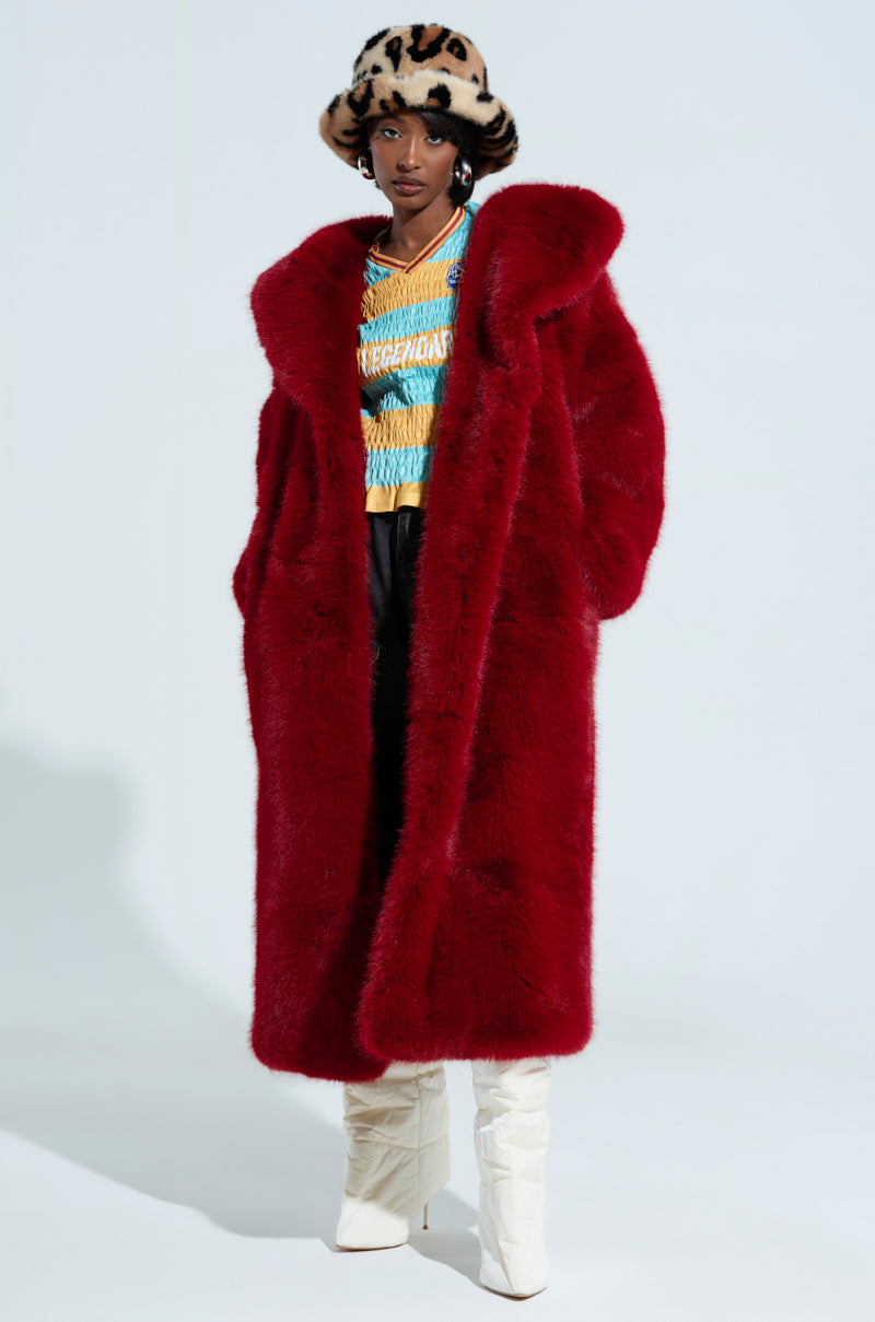 A person wearing the MANCHESTER HOODED FAUX FUR IN WINE, along with a colorful knitted top, black pants, a leopard-print hat, and tall white boots stands against a plain background. Their hands are in their coat pockets, and they have a confident expression.