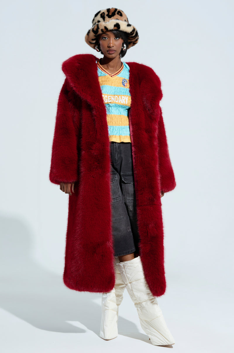 A person dons the Manchester Hooded Faux Fur in a plush wine hue over a colorful shirt emblazoned with "LEGENDARY." They accessorize with a patterned hat, dark pants, and white knee-high boots, while the plain light-colored background enhances their vibrant ensemble.