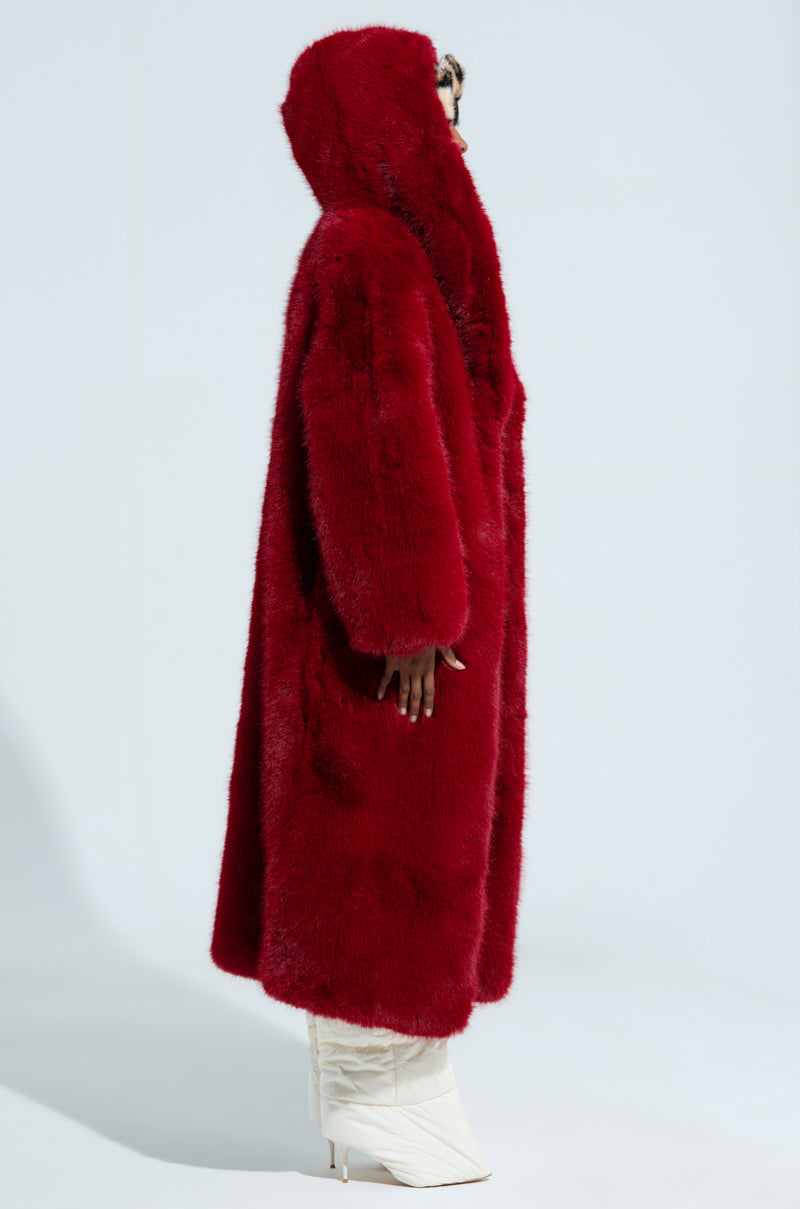 Dressed in the MANCHESTER HOODED FAUX FUR IN WINE, an individual is seen from the side, their face half-hidden. They complement this elegant look with sleek white pointed-heeled boots set against a simple, light-colored backdrop.
