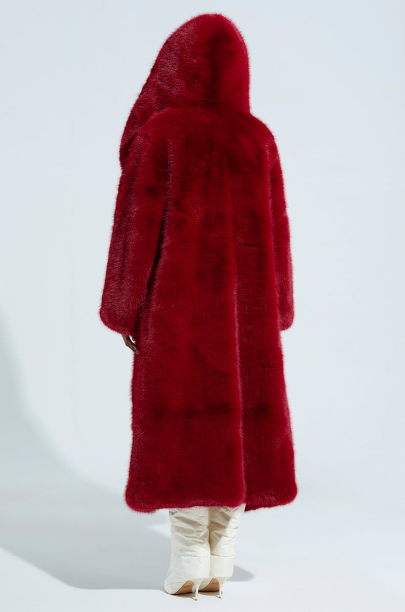 Dressed in the luxurious MANCHESTER Hooded Faux Fur coat in a rich wine color, an individual stands with their back to the camera. They pair it with sleek white, pointed-toe boots against a minimalist light gray backdrop.