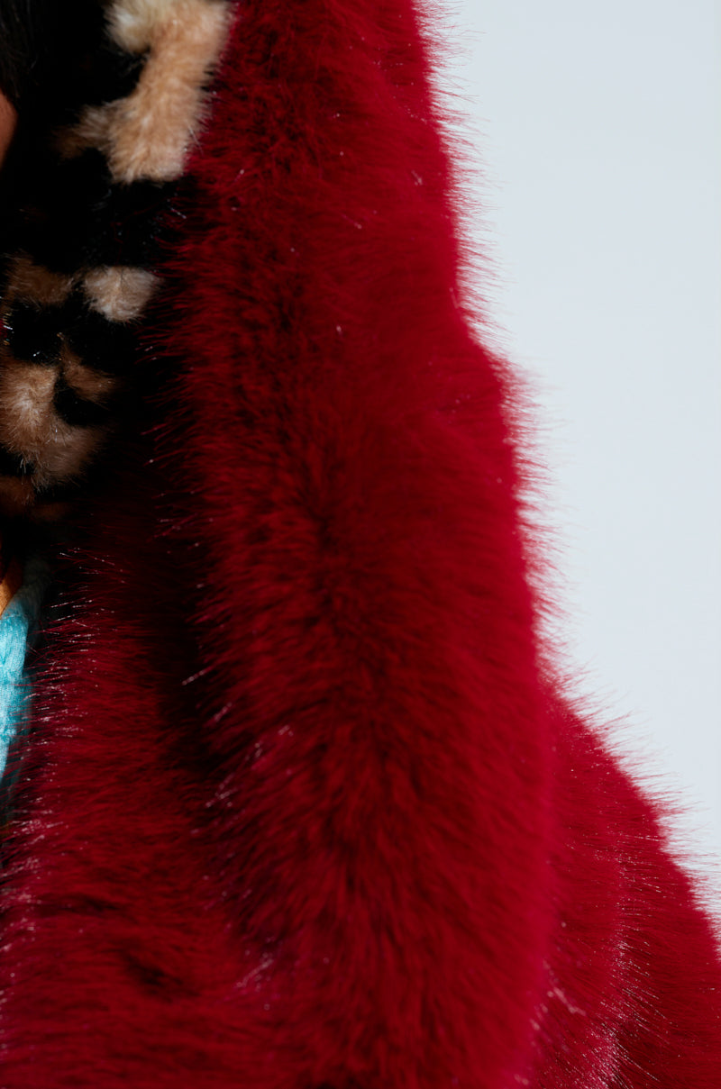 A close-up of the vibrant MANCHESTER HOODED FAUX FUR IN WINE showcases its luxuriously soft and plush texture. The edge features a hint of leopard print fabric, providing a striking contrast to the rich wine red. The background is a minimalist, pale gray.