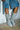 A person wearing AZALEA WANG MANSI SILVER BUCKLE DETAIL BOOTS, which feature a lace-up vamp and chunky block heel, paired with denim shorts adorned with decorative buckles and straps. They are standing on a concrete floor next to a white, metal lattice-pattern wall.