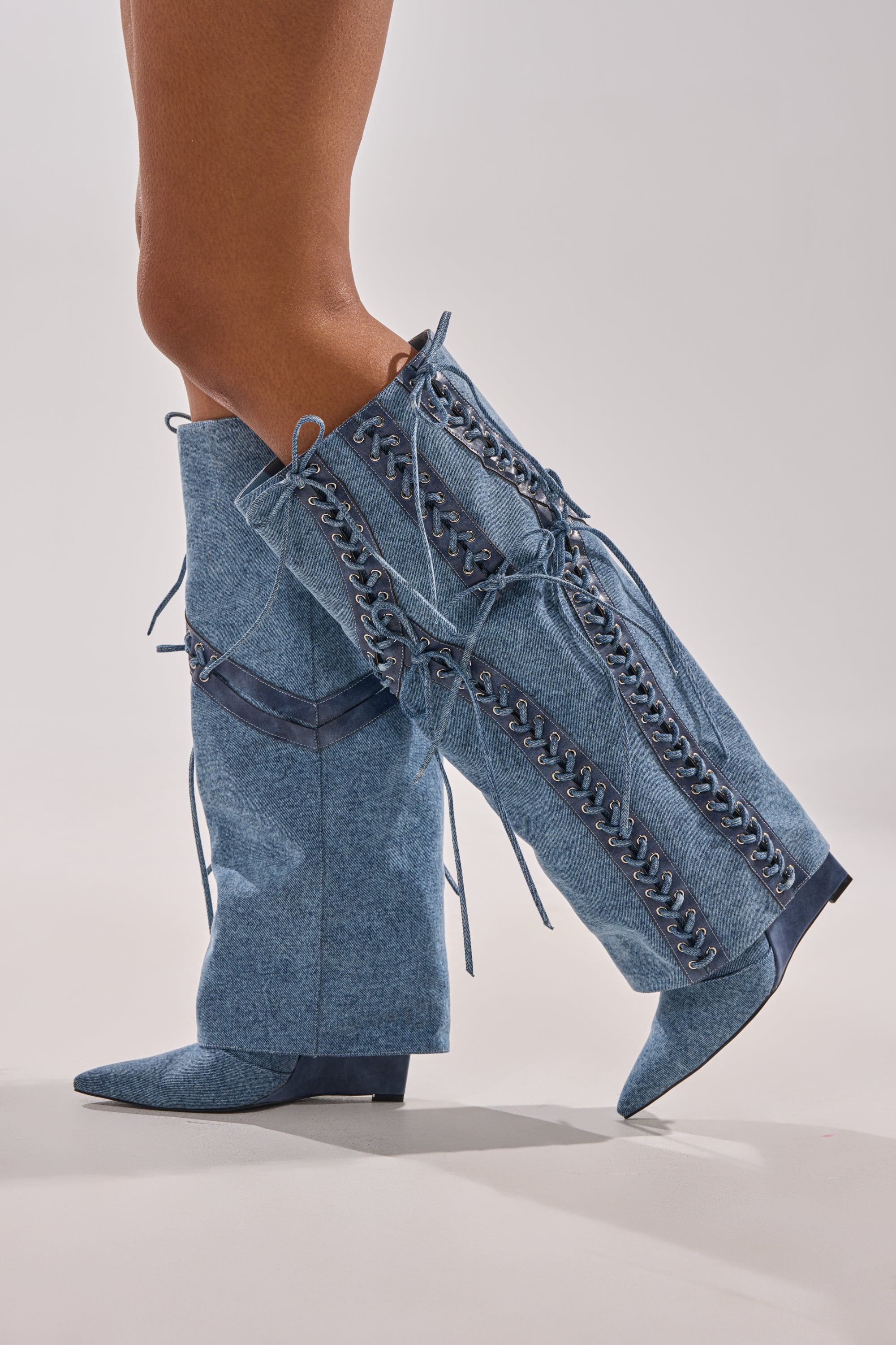 A close-up side view of the AKIRA GOLD MANTIGNA DENIM WEDGE BOOT, featuring knee-high corset lacing, pointy denim design, dark blue ribbons, and low heels against a neutral background.