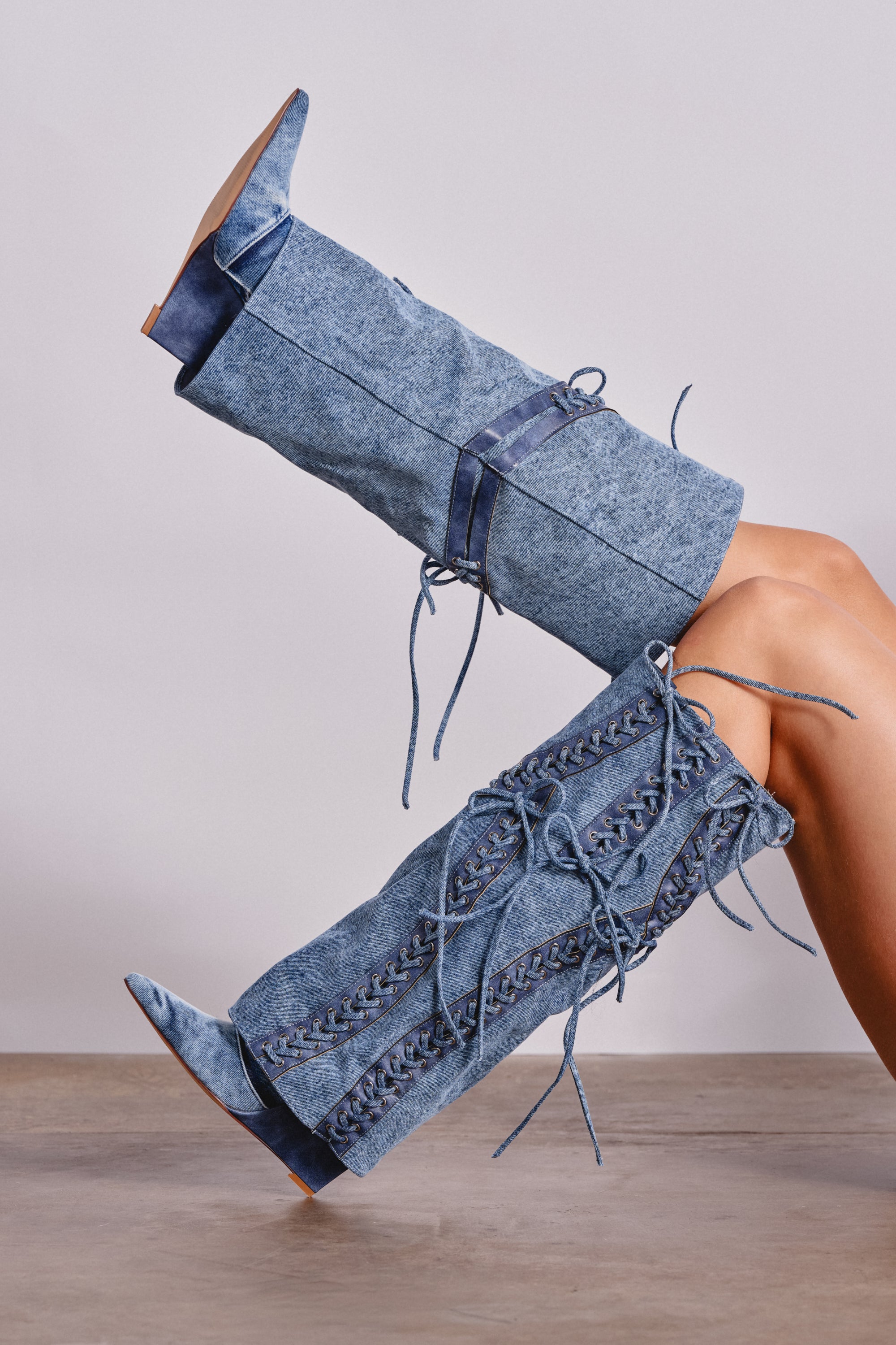 Legs crossed at the knee, wearing AKIRA GOLD MANTIGNA DENIM WEDGE BOOT—tall, light blue lace-up denim knee-high boots with high heels and pointed toes; neutral background with wooden floor.