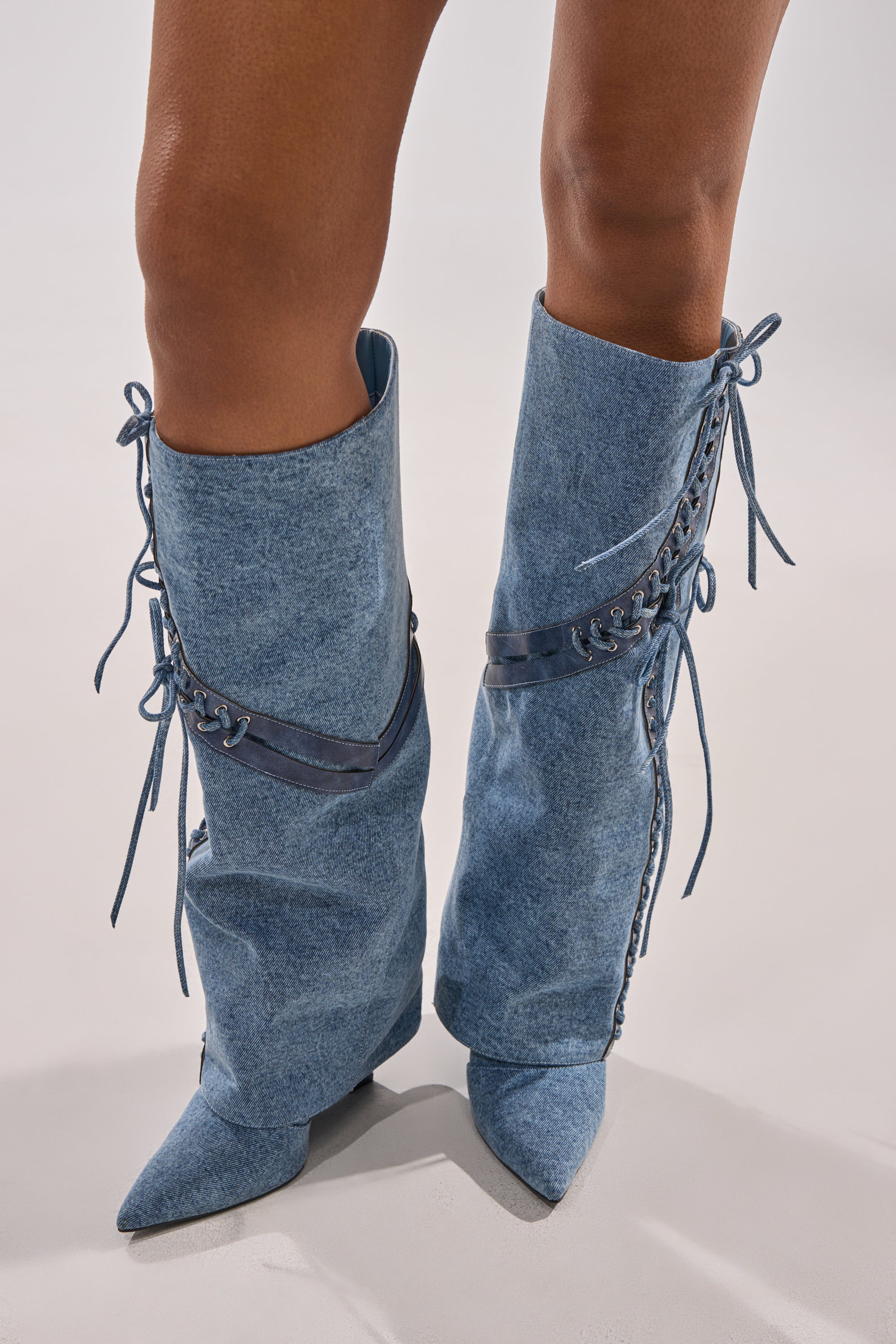 A person stands on a light surface wearing the AKIRA GOLD MANTIGNA DENIM WEDGE BOOTS, featuring knee-high length, pointed toes, corset-style lacing, and dark blue leather accents.