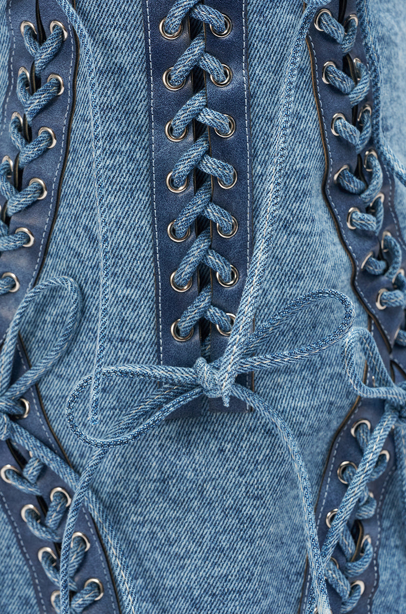 Close-up of AKIRA GOLD MANTIGNA DENIM WEDGE BOOT shows blue denim fabric with corset-style lacing through metal eyelets, tied neatly at the center. The detailed stitching and laces spotlight this stylish wedge boot’s design.