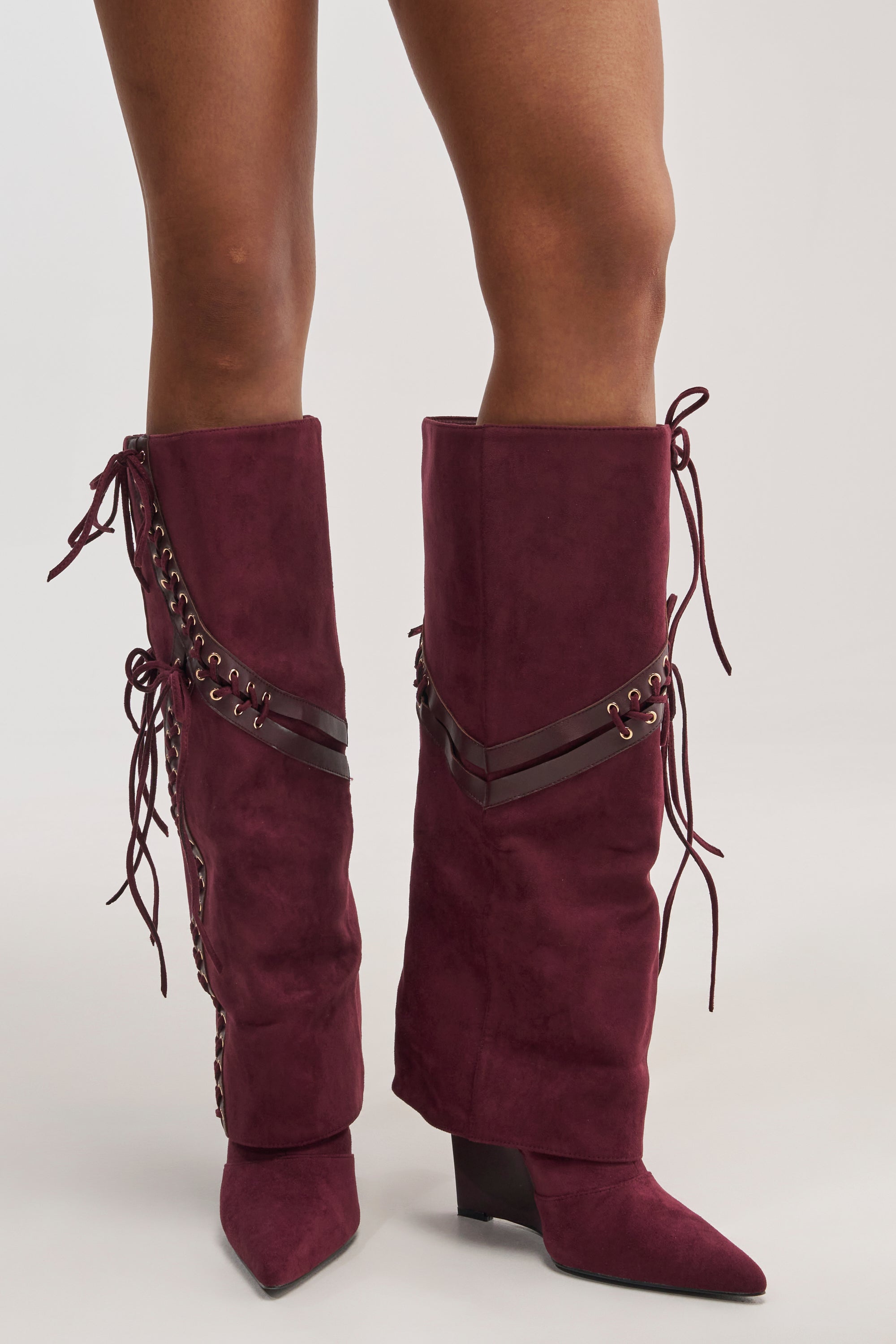 A model wears the AZALEA WANG MAGENTA PLUM FAUX SUEDE WEDGE BOOTS—knee-high wedge boots in a rich magenta plum faux suede, featuring leather strap detailing, gold-toned eyelets, and long decorative lace-up ties running along the sides and calf.