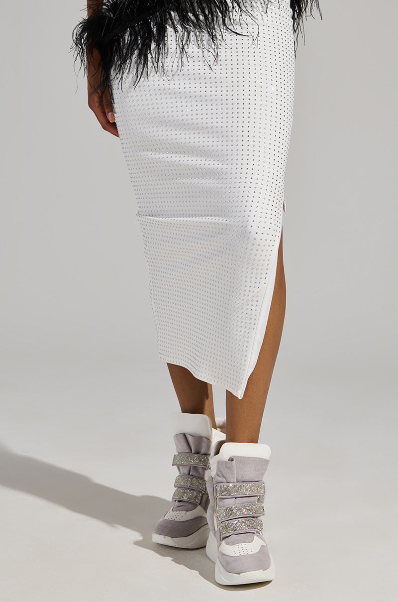 A person is depicted from behind wearing a knee-length white skirt with a textured pattern and a fuzzy black waistband. They are sporting gray Maranto White Wedge Sneakers, featuring rhinestone-embellished velcro foot straps. The background is plain gray.