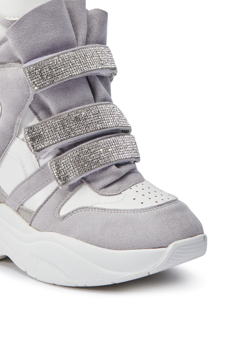 A detailed view of the MARANTO WHITE WEDGE SNEAKER highlights its thick sole and stylish design. This shoe features light grey faux suede panels paired with rhinestone-embellished velcro foot straps, and the perforated white toe area enhances both breathability and style.