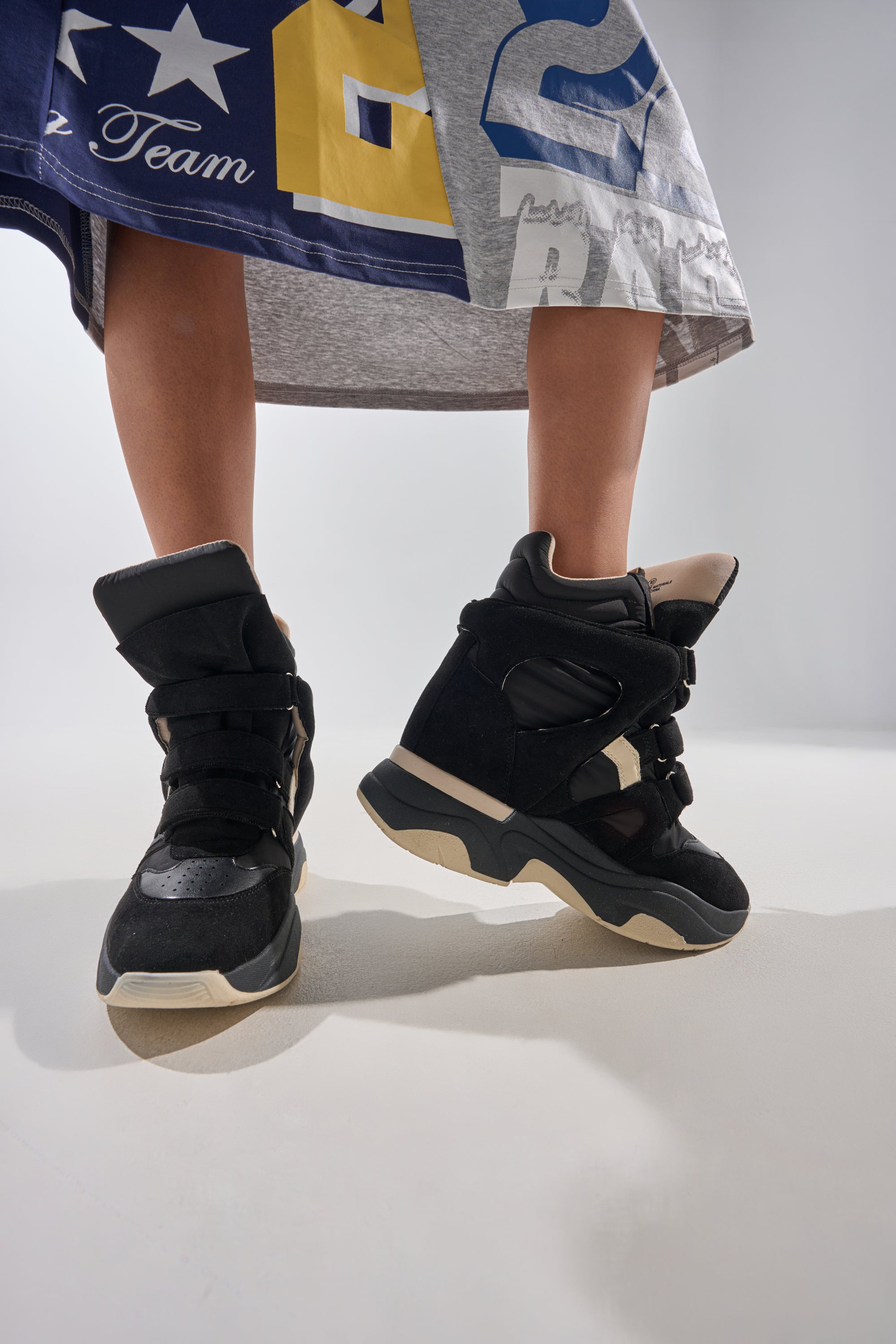 A person stands on a white surface, legs slightly apart and toes out, wearing MARANTO CHUNKY BLACK SNEAKER with a gray skirt featuring blue and yellow designs. 0