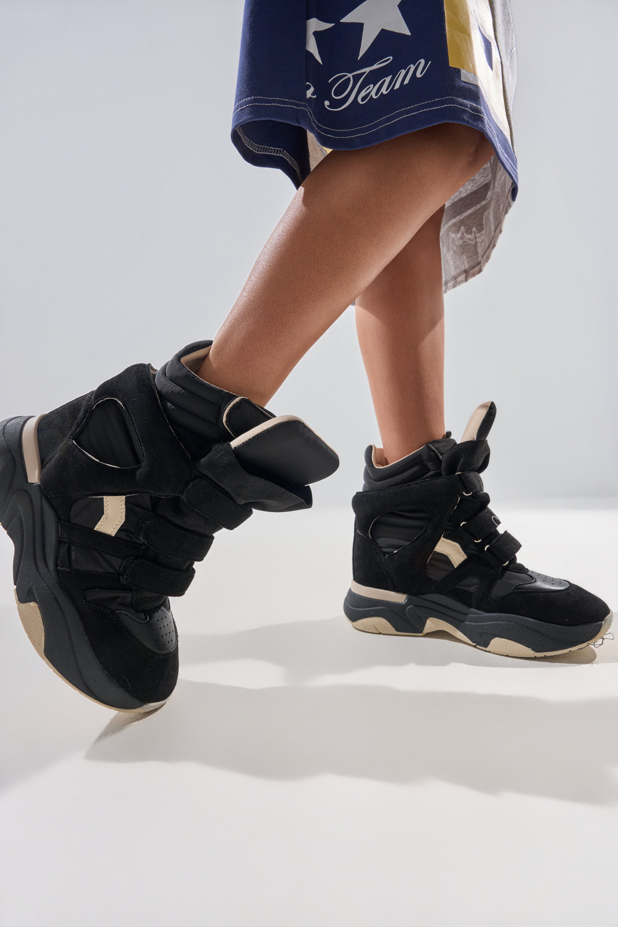 A person wears the MARANTO CHUNKY BLACK SNEAKER with a wedge heel, standing with one foot raised. They pair it with a blue and white skirt or shorts decorated with yellow stars and text. 1