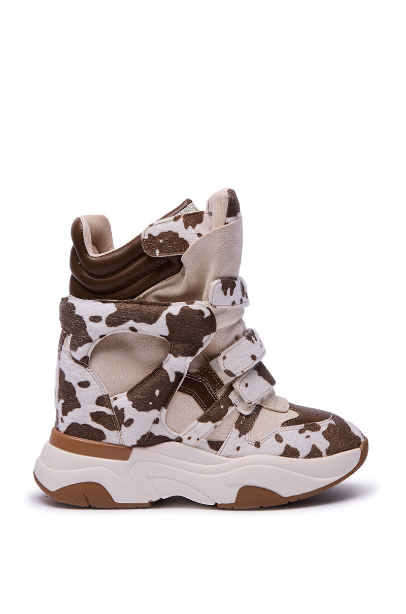The MARANTO COW COLOR BLOCKED WEDGE SNEAKER features a cow print, brown suede accents, chunky white sole, and velcro straps—making it a playful high-top for color-blocked sneaker fans. Shown against a white background.