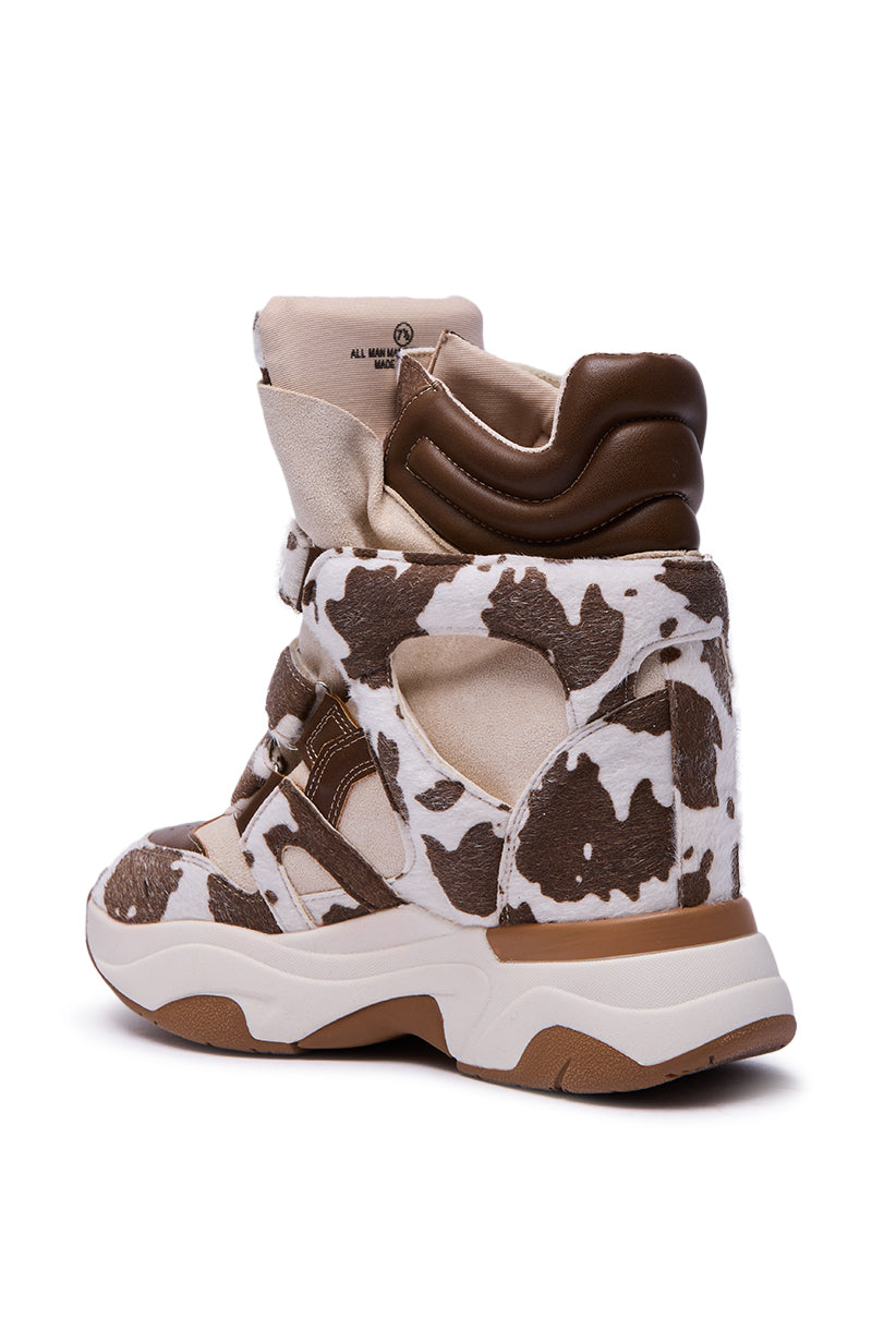 The MARANTO COW COLOR BLOCKED WEDGE SNEAKER features a high-top design with brown and white cow-print, beige accents, thick white sole, hidden wedge heel, and a padded brown ankle collar, shown from a side-rear angle.