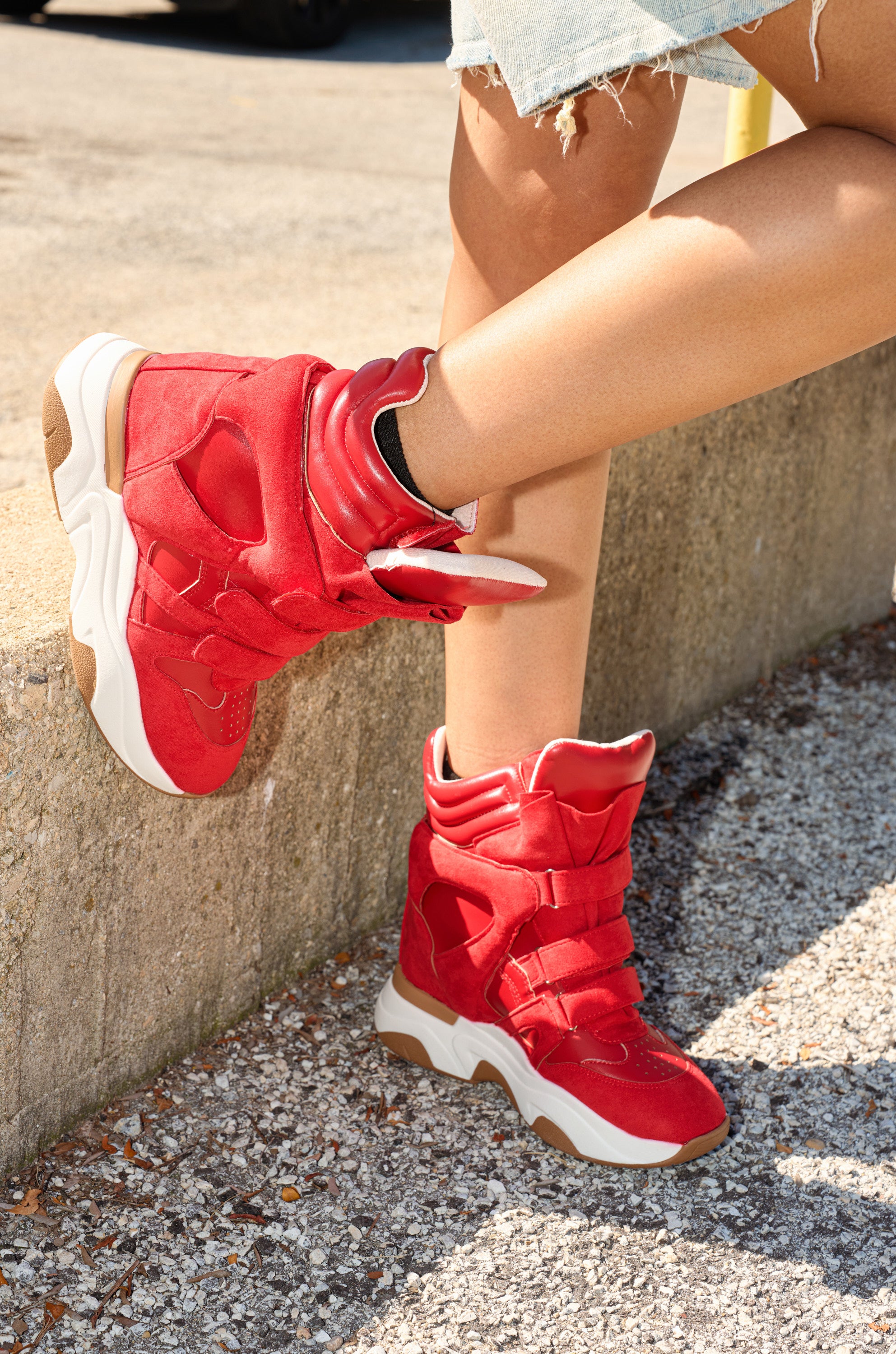 Someone sitting outdoors on a concrete ledge, one leg crossed over the other, wearing ripped denim shorts and MARANTO RED WEDGE SNEAKER with thick soles.