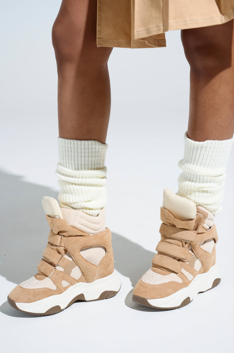 A close-up showcases a person wearing MARANTO TAUPE WEDGE SNEAKERS with thick laces and white ribbed socks. The faux leather shoes feature a chunky sole, highlighting their stylish design against a neutral backdrop, complemented by a tan skirt.