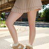 A person in a pleated beige mini skirt and MARANTO TAUPE WEDGE SNEAKER stands on a sunlit city street, with an elevated train track and brick buildings in the background.