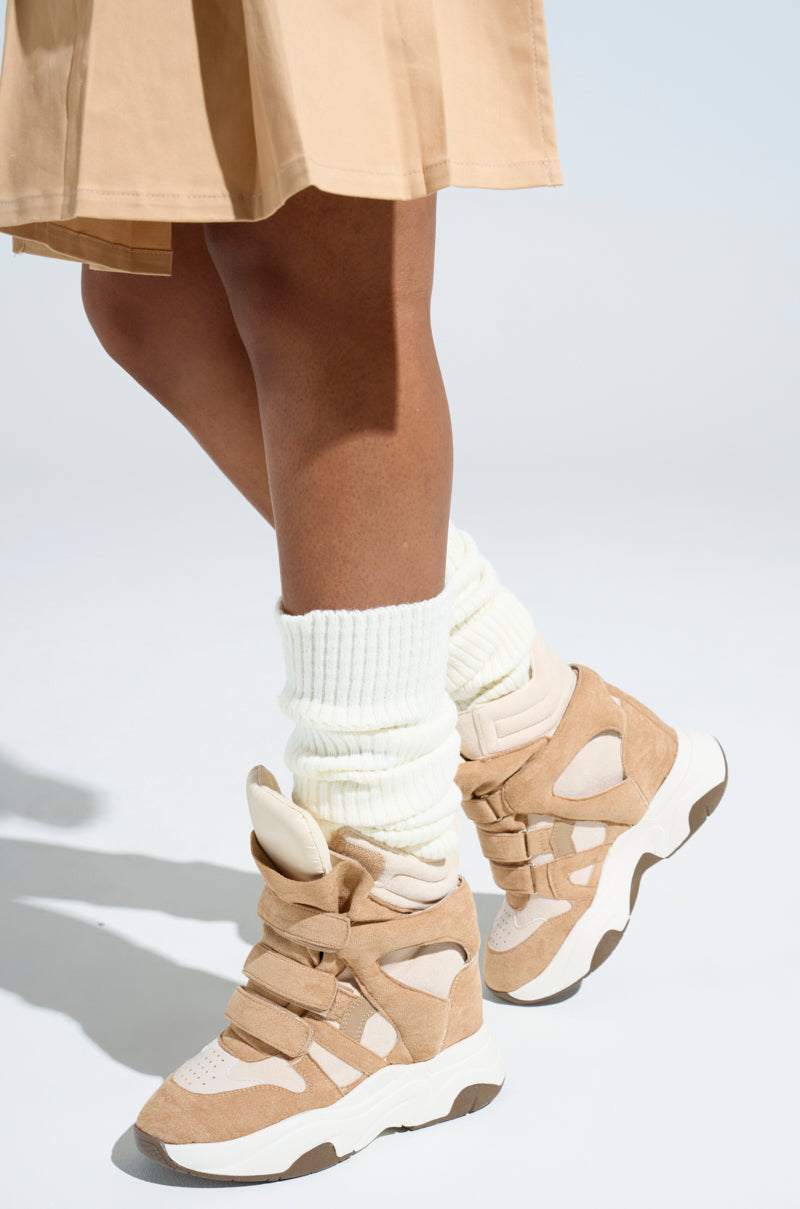 Someone wears MARANTO TAUPE WEDGE SNEAKER featuring chunky soles and a concealed wedge heel, paired with white leg warmers and a beige skirt against a neutral backdrop.