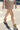 Wearing a beige pleated mini skirt, white top, metallic bracelets, and MARANTO TAUPE WEDGE SNEAKER with a concealed wedge heel, someone walks across a sunlit street crosswalk.